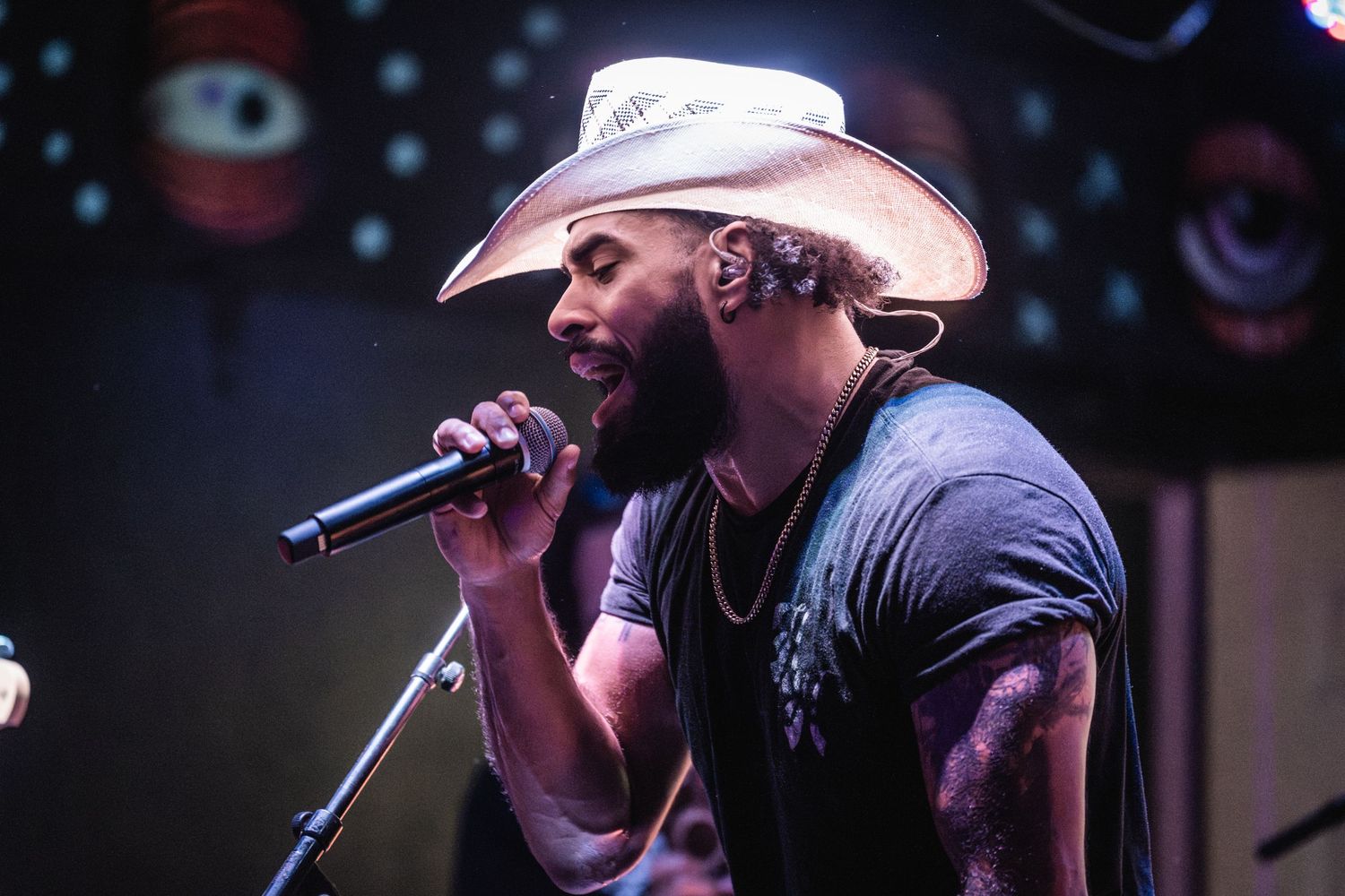 Performer in cowboy hat and dark shirt holding microphone on stage with colorful lighting effects.