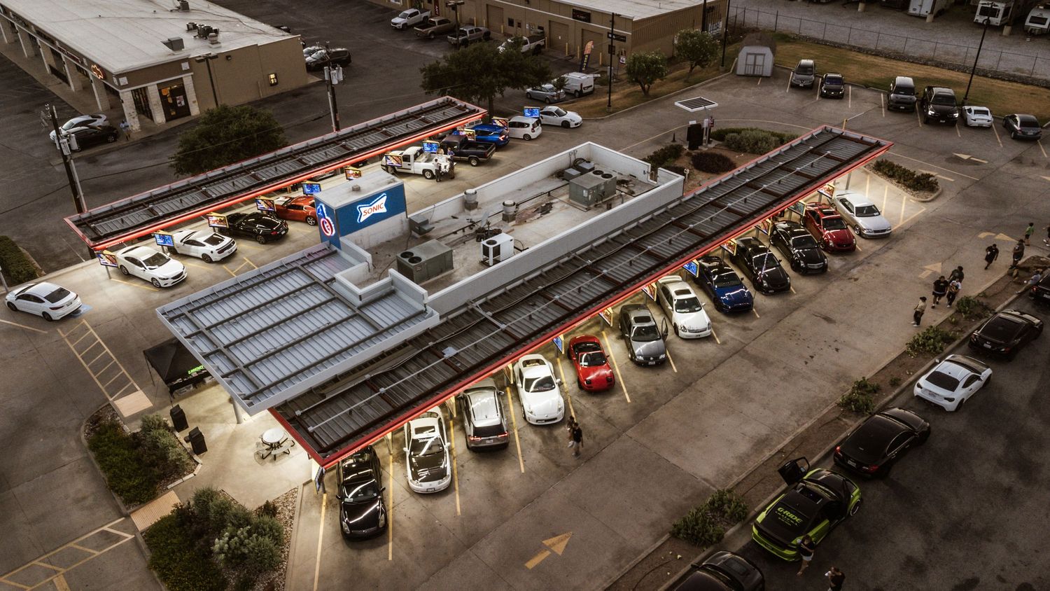 Aerial view of a car meet at night showing rows of vehicles parked under bright lights.