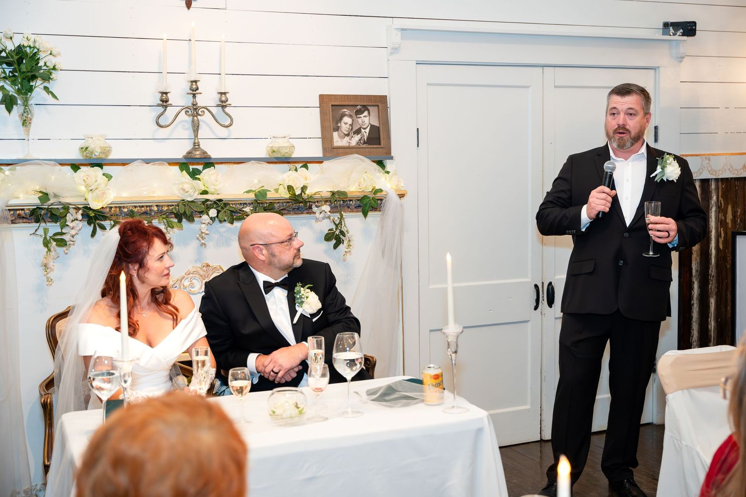 Wedding reception speech being given at a white decorated venue with guests seated at tables.