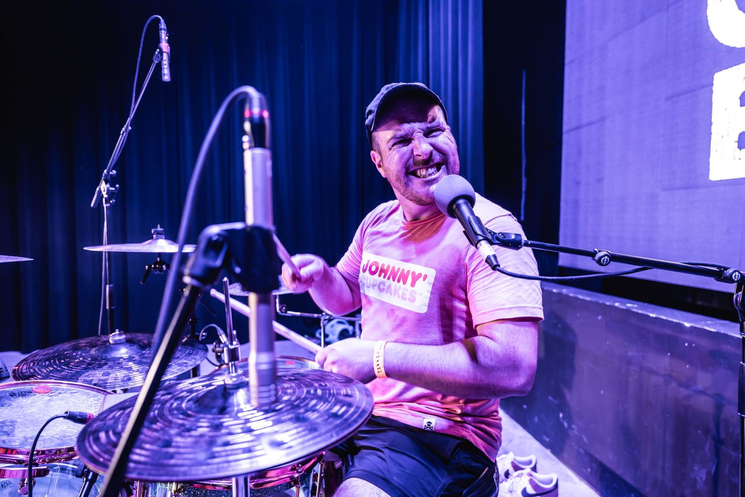 Drummer performing enthusiastically behind drum kit on illuminated stage.