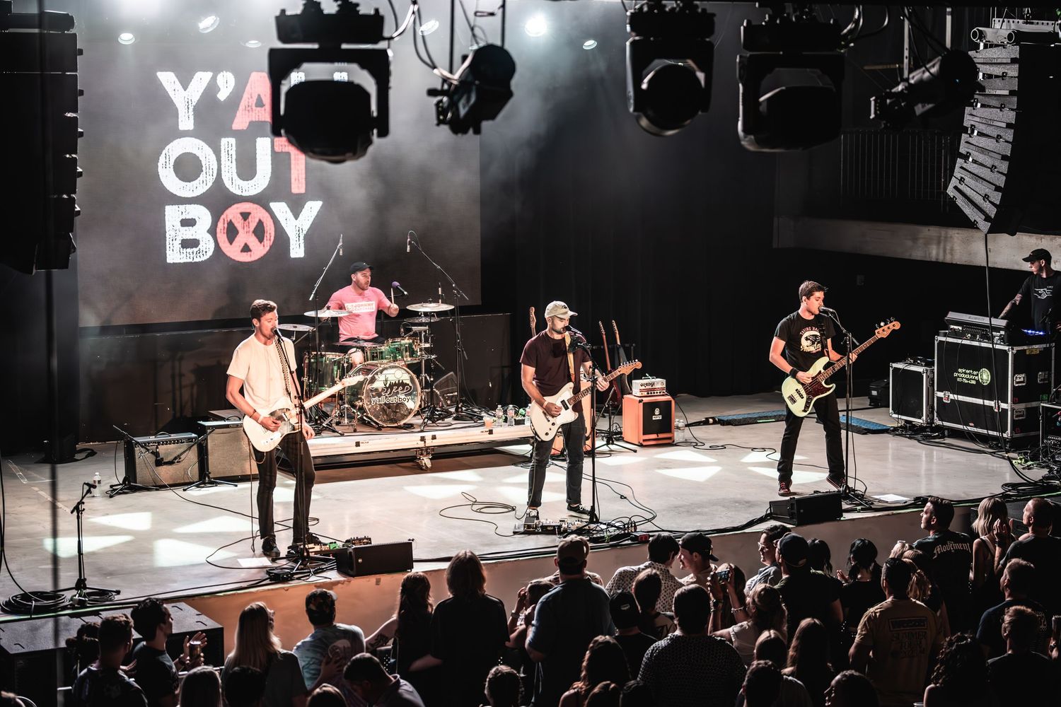 Rock band performs on brightly lit stage with Y'ALL OUT BOY logo displayed behind them.