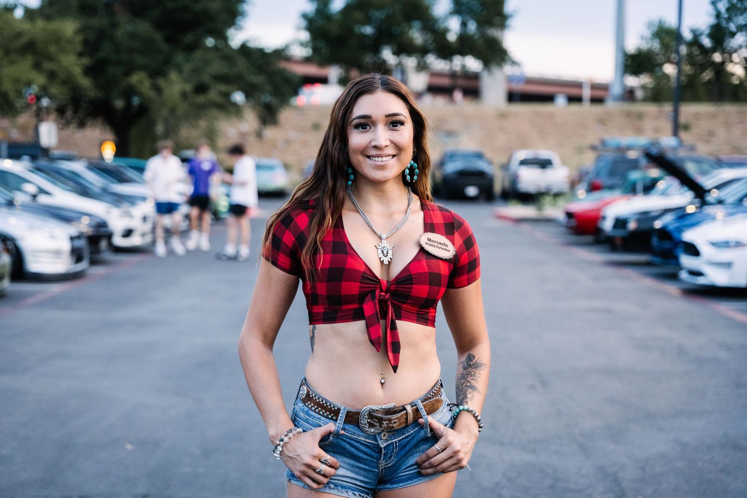 Person in red plaid crop top and denim shorts posing in parking lot with cars in background.