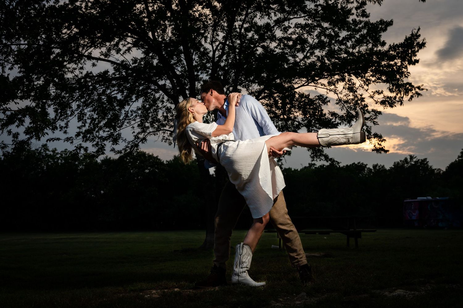 Romantic couple in formal attire shares an intimate dance dip at sunset under a tree in a park setting.