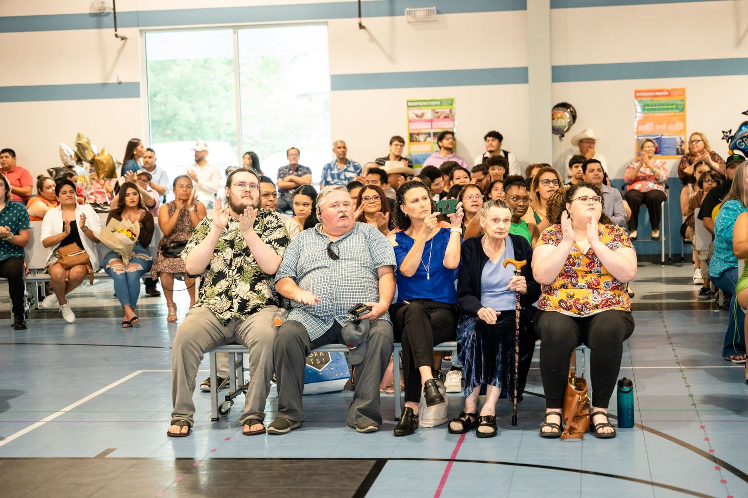 Large audience gathered in a gymnasium-style room with blue trim watching an event.