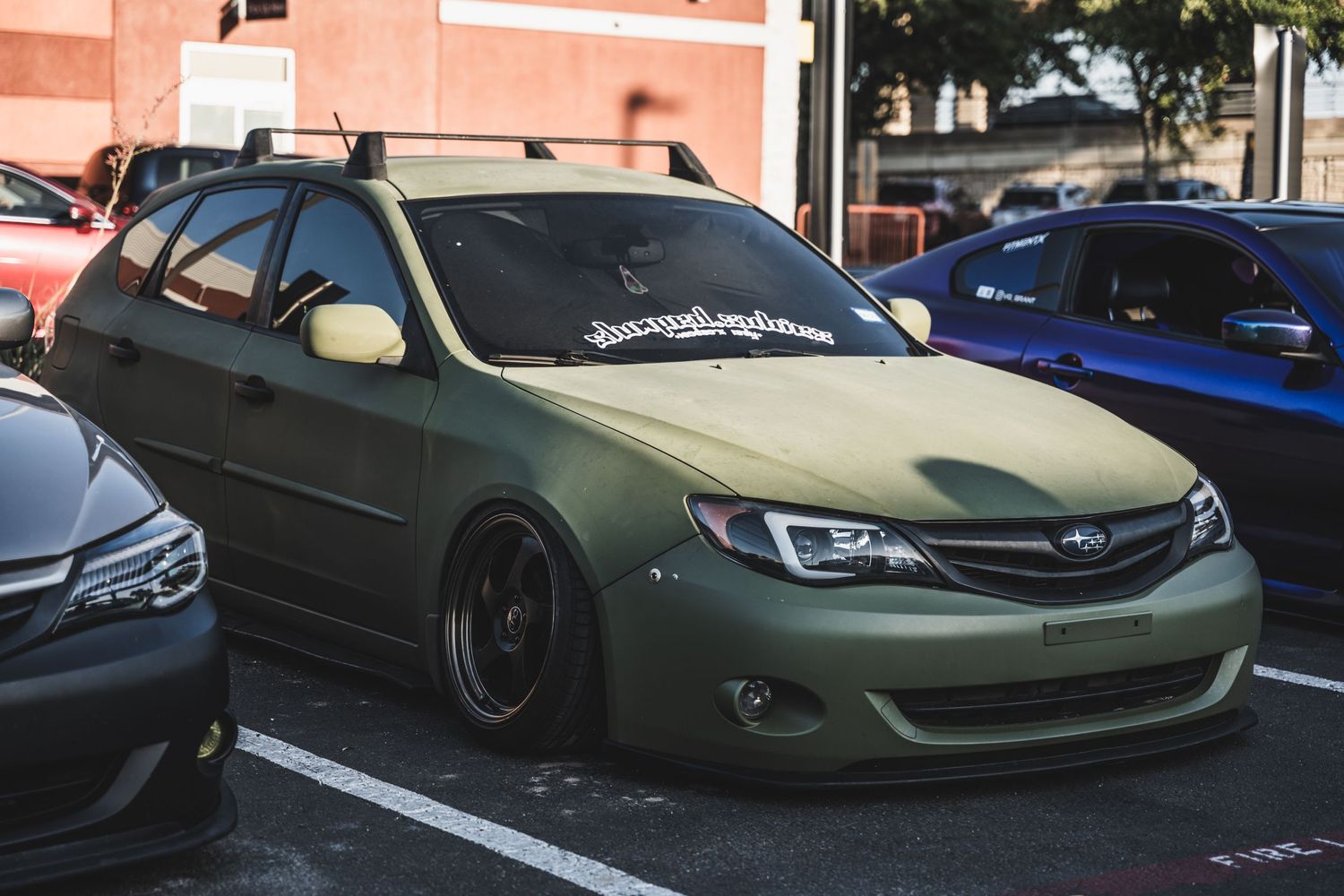 Matte olive green modified Subaru Impreza with lowered suspension in a parking lot.