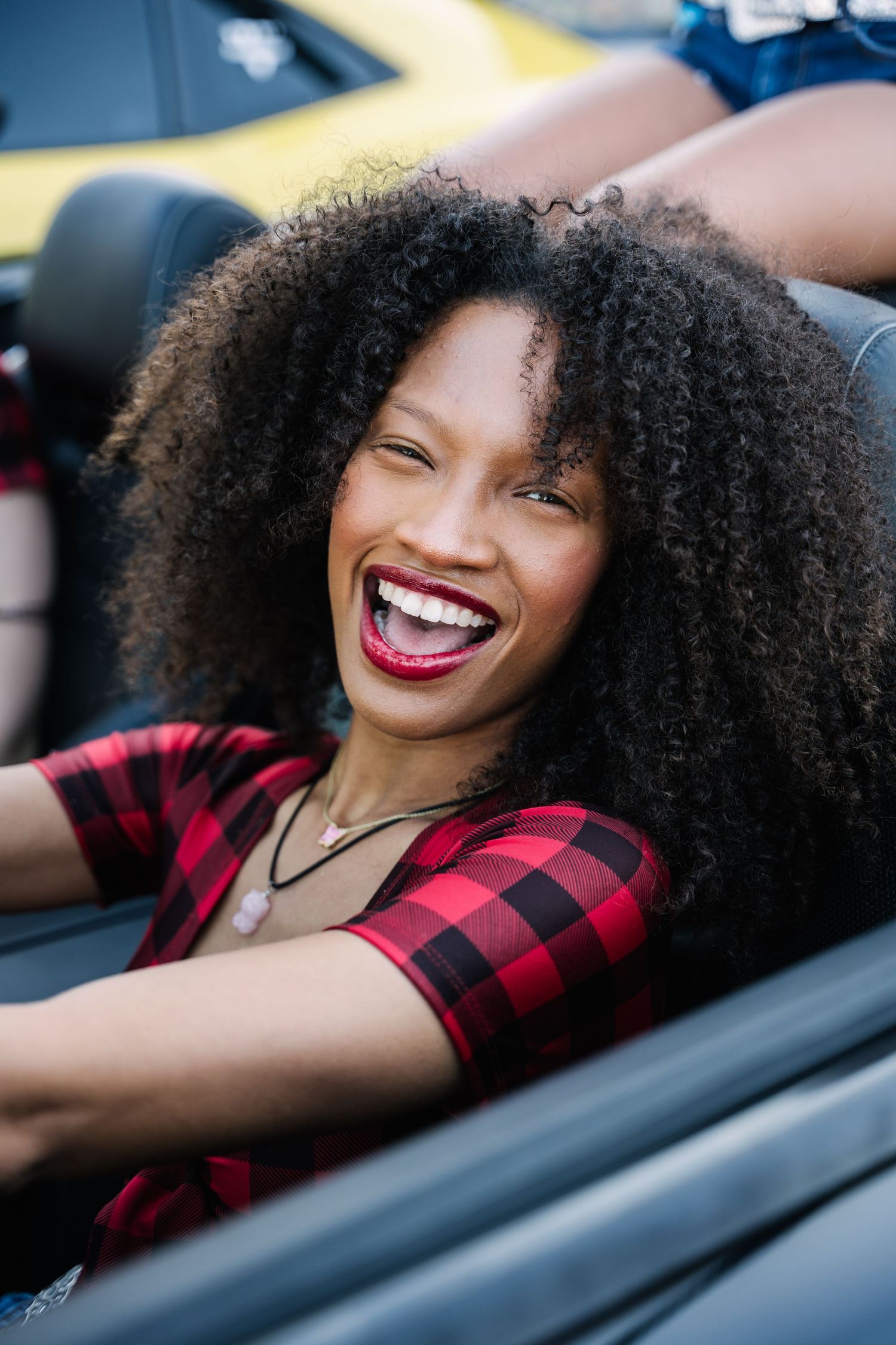 A model with natural curly hair smiles while sitting in a convertible car.