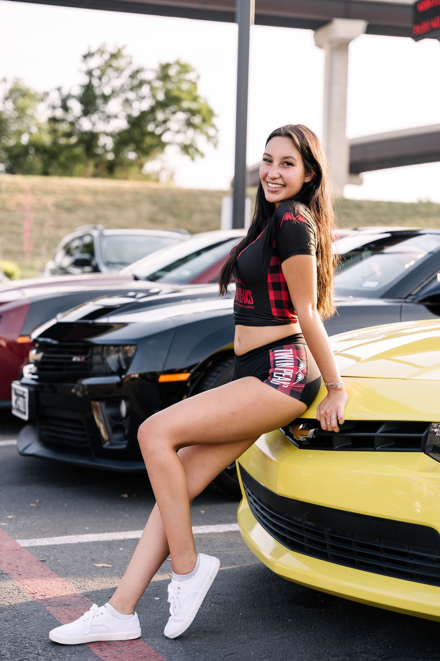 Young woman in casual attire posing by yellow sports car in parking lot.