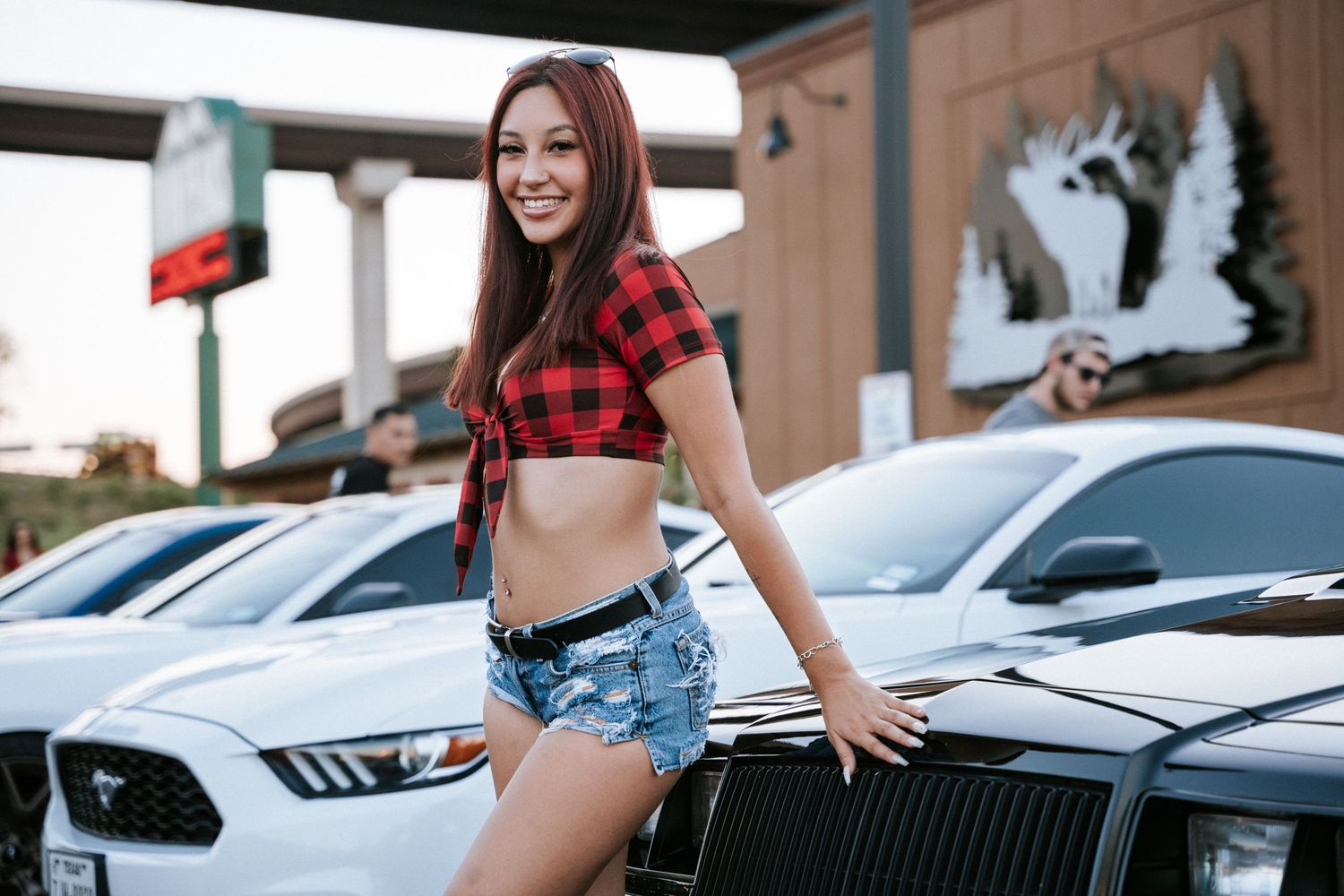 Model in red plaid top posing beside vintage sports car at automotive event.
