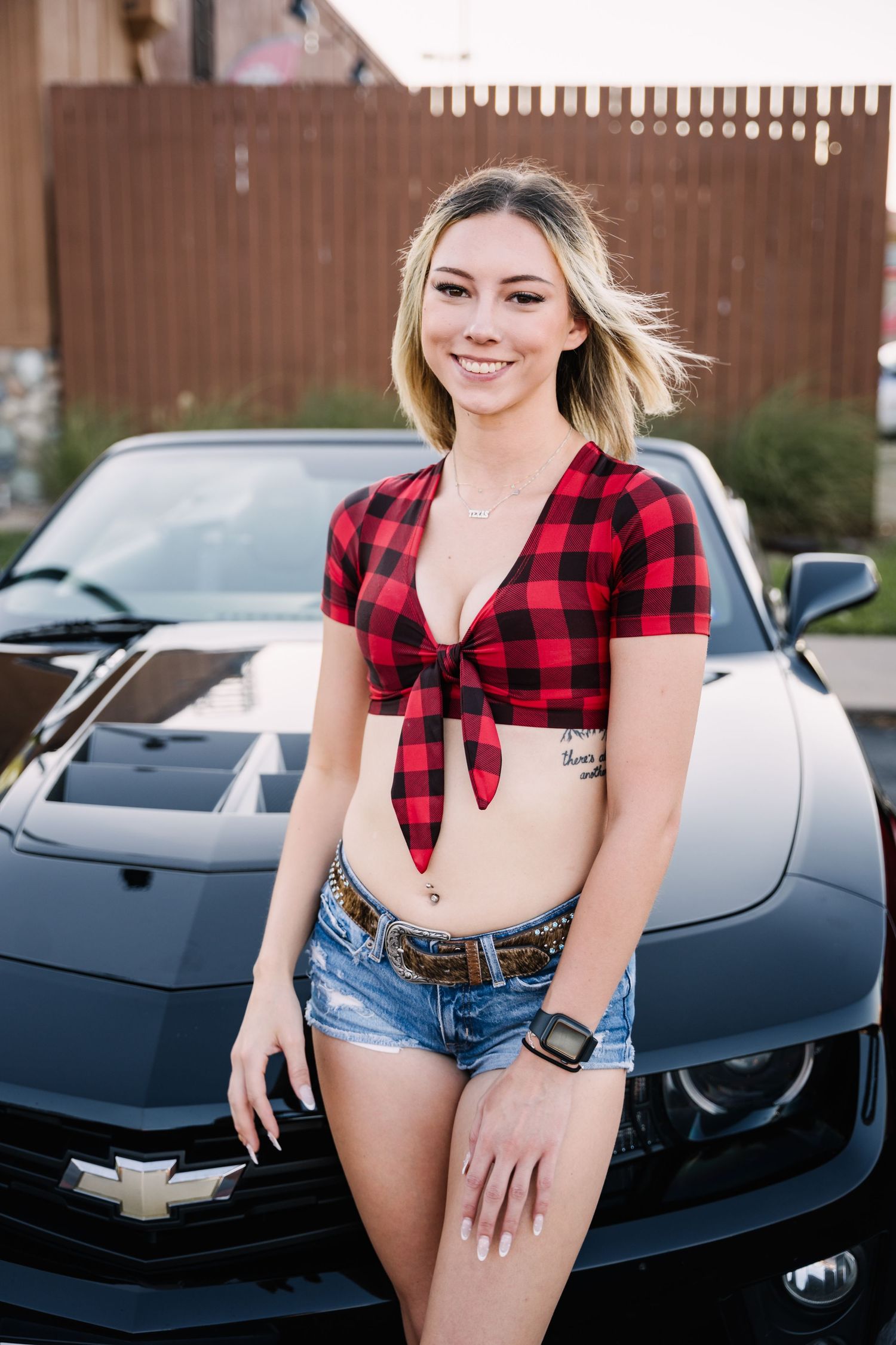 Person in red plaid crop top and denim shorts poses in front of black Chevrolet.