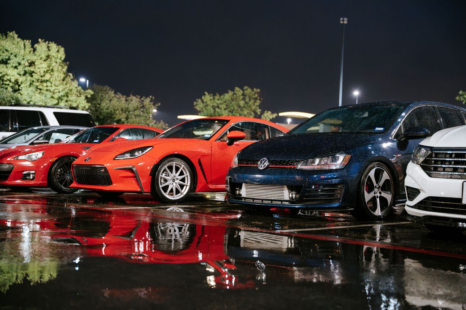 Line of colorful modified sports cars reflecting in wet pavement at a car show.