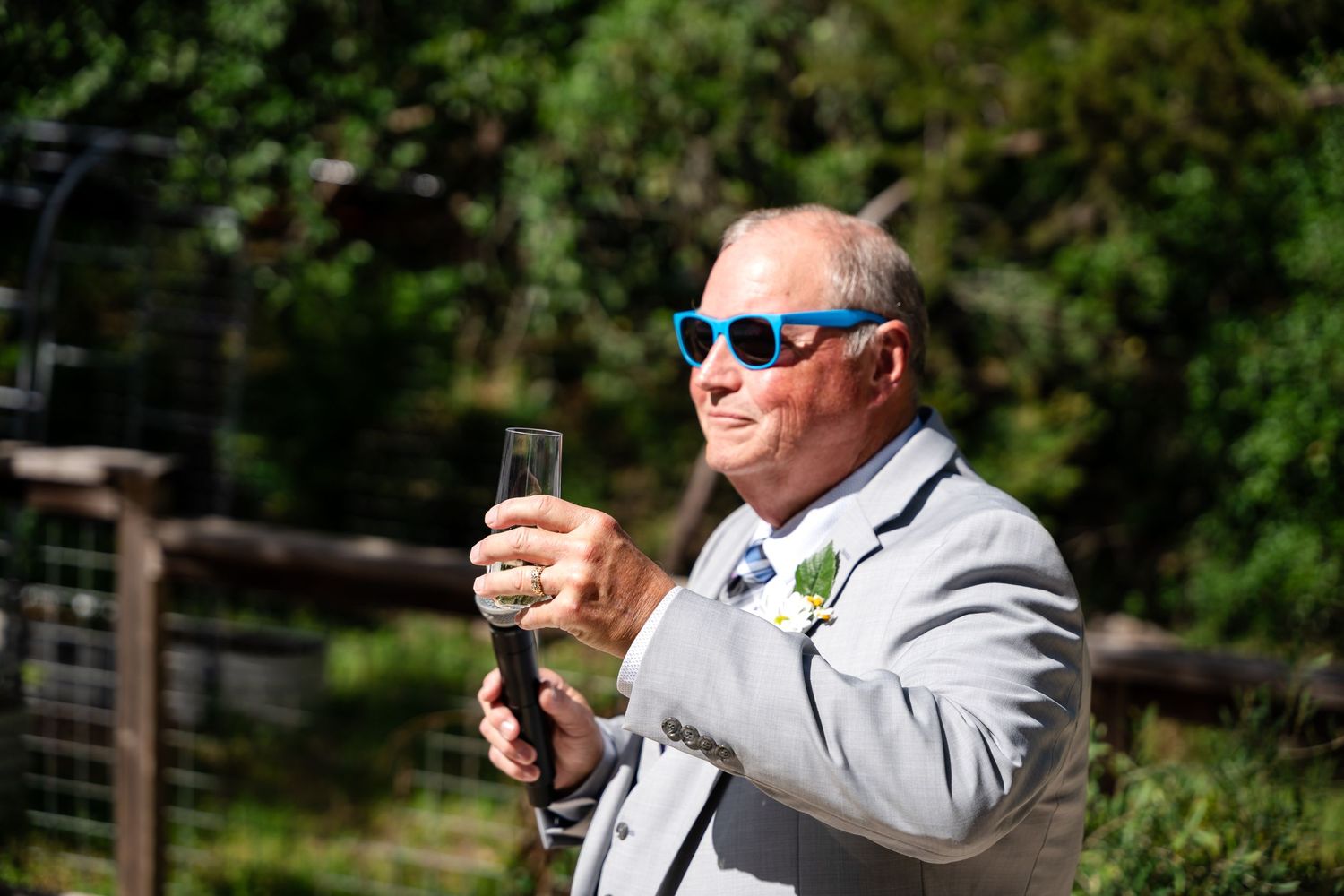 A guest in a grey suit raises a champagne glass at an outdoor celebration.