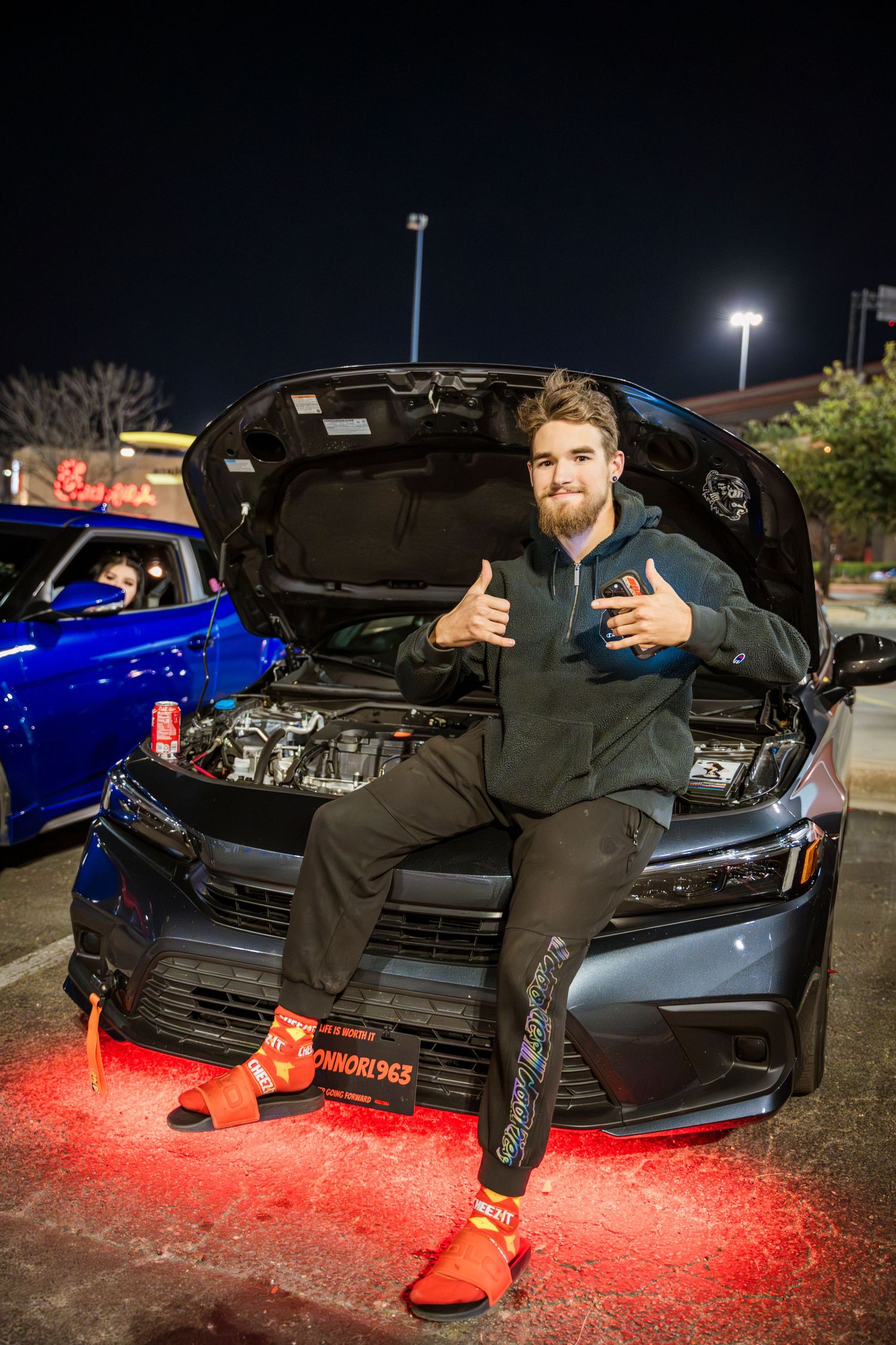 Car enthusiast gives thumbs up while sitting on hood of modified vehicle with underbody lights.