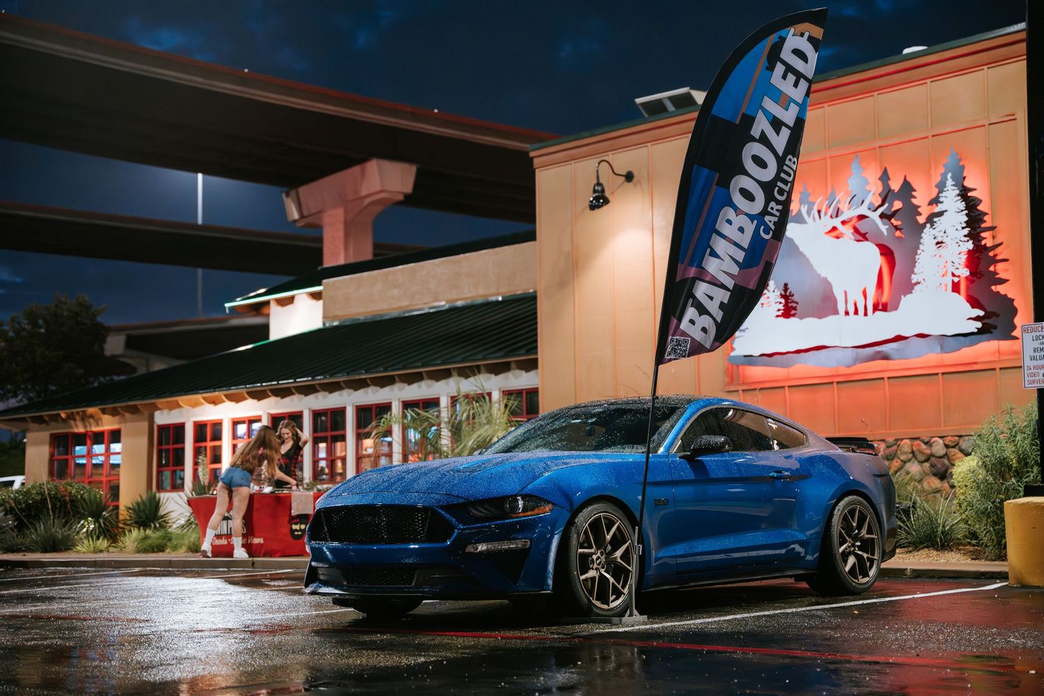 Blue Ford Mustang parked in front of a retail building at night with wet pavement.
