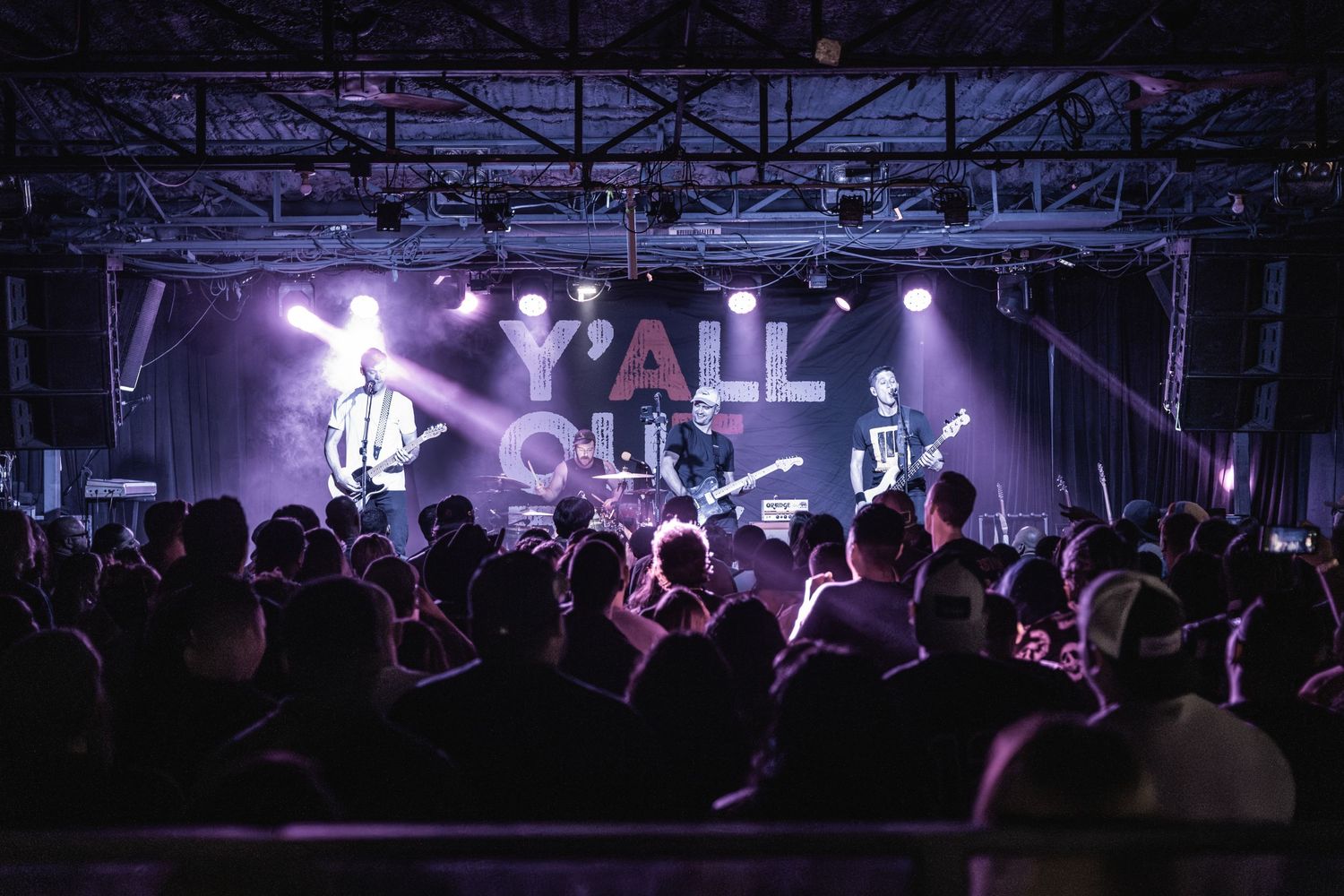 Concert venue with band performing under purple stage lights as crowd watches below.