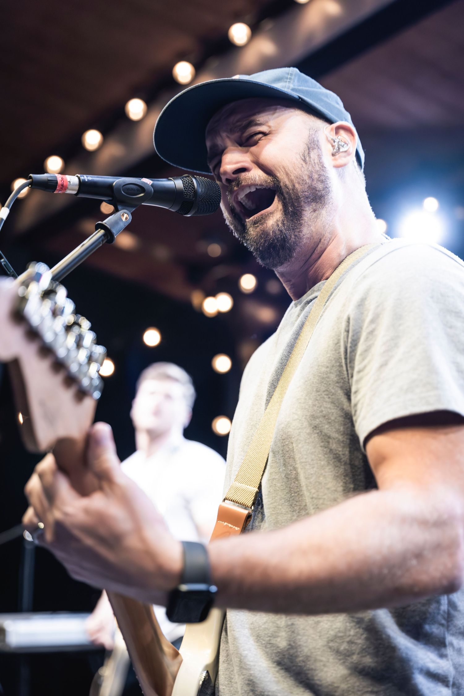 Musician wearing baseball cap performs with guitar and sings into microphone on illuminated stage.