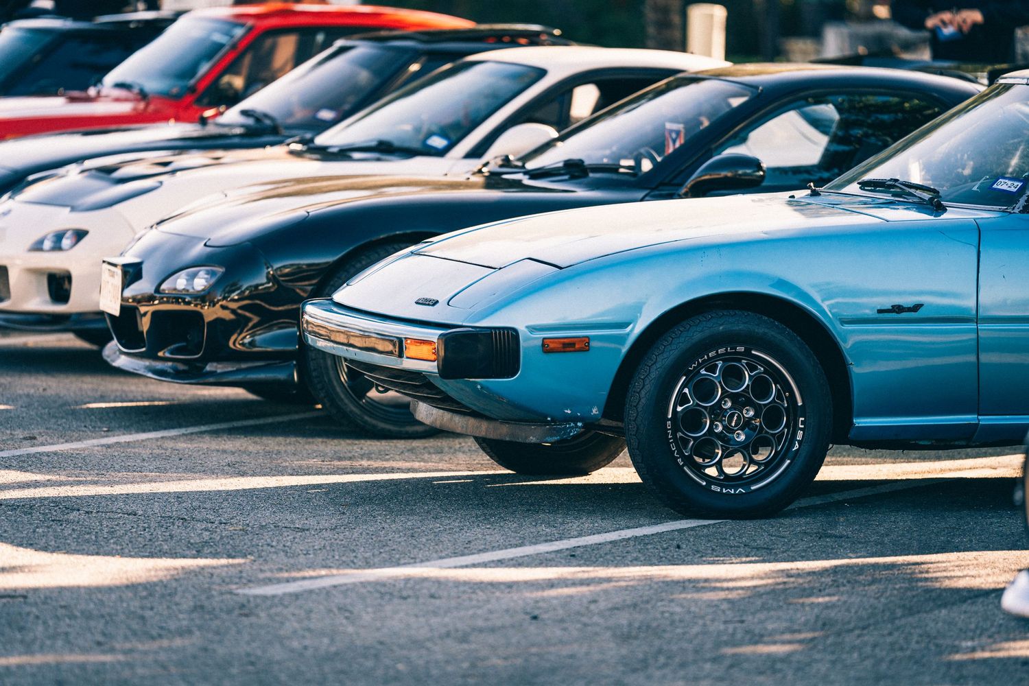 Row of vintage Japanese sports cars including a light blue Mazda RX-7 in a parking lot.