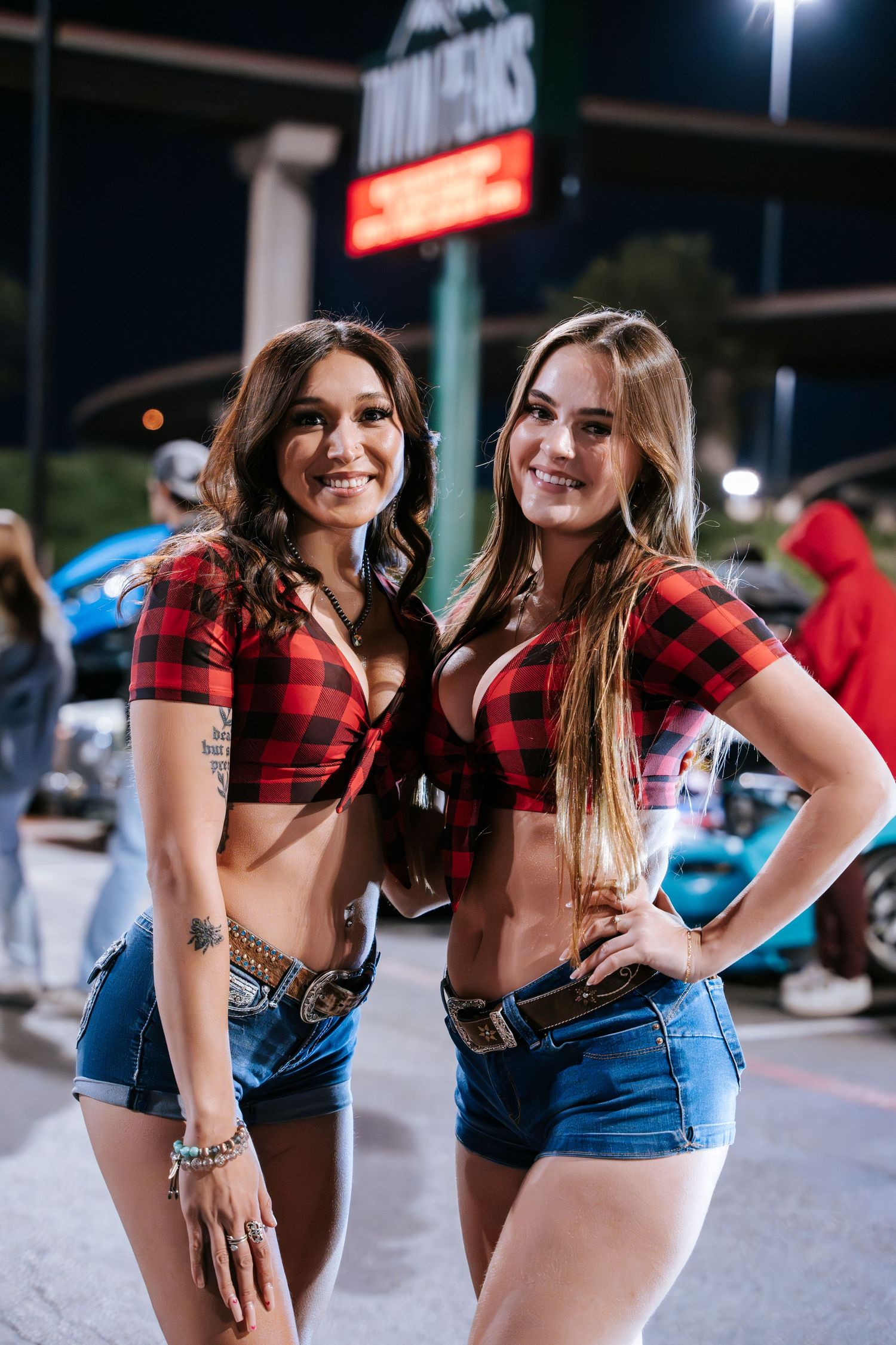 Two women pose in matching red plaid crop tops and denim shorts at a nighttime gathering.