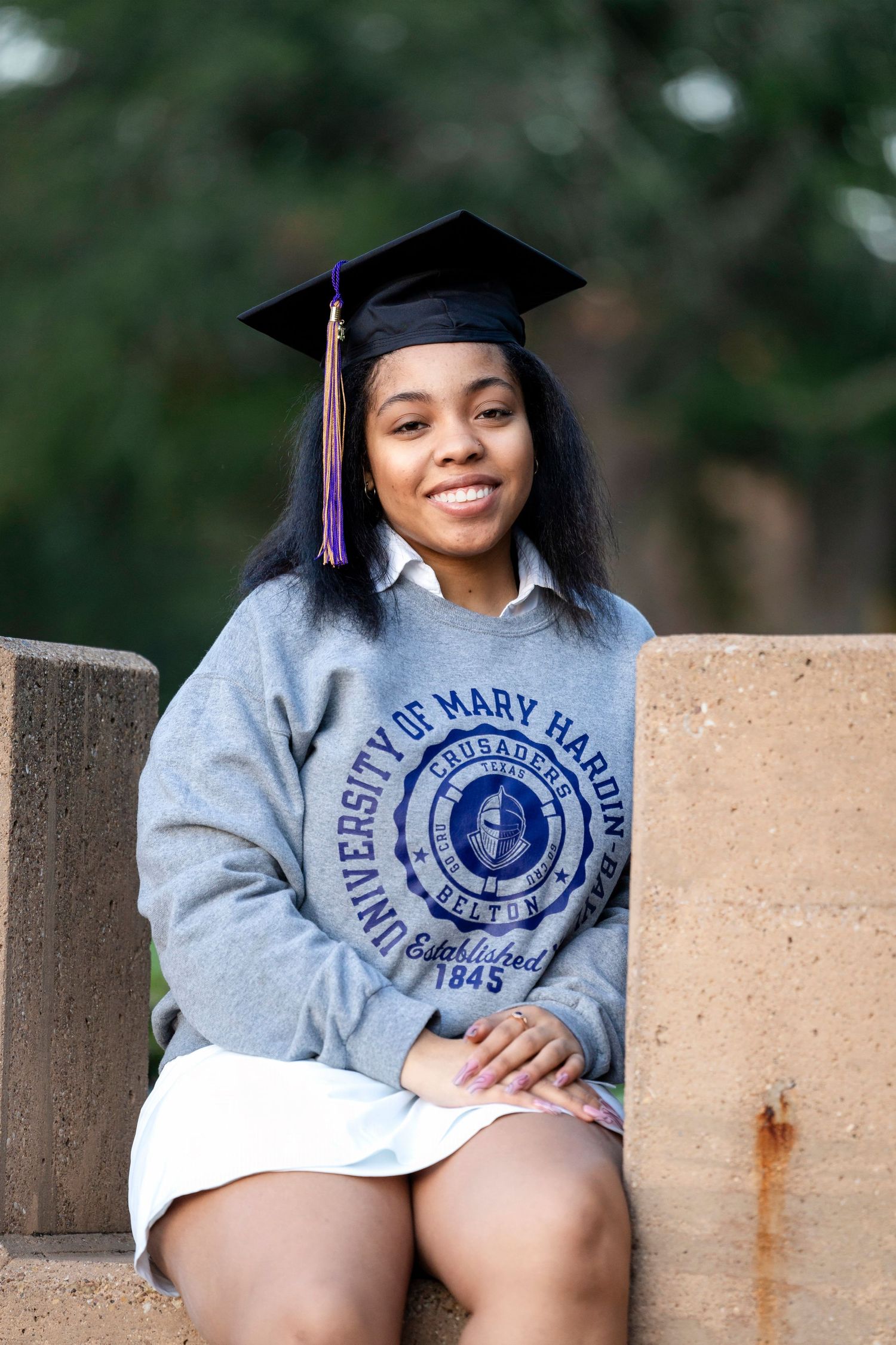 Graduate in cap and University of Mary Hardin-Baylor sweatshirt seated on concrete structure.