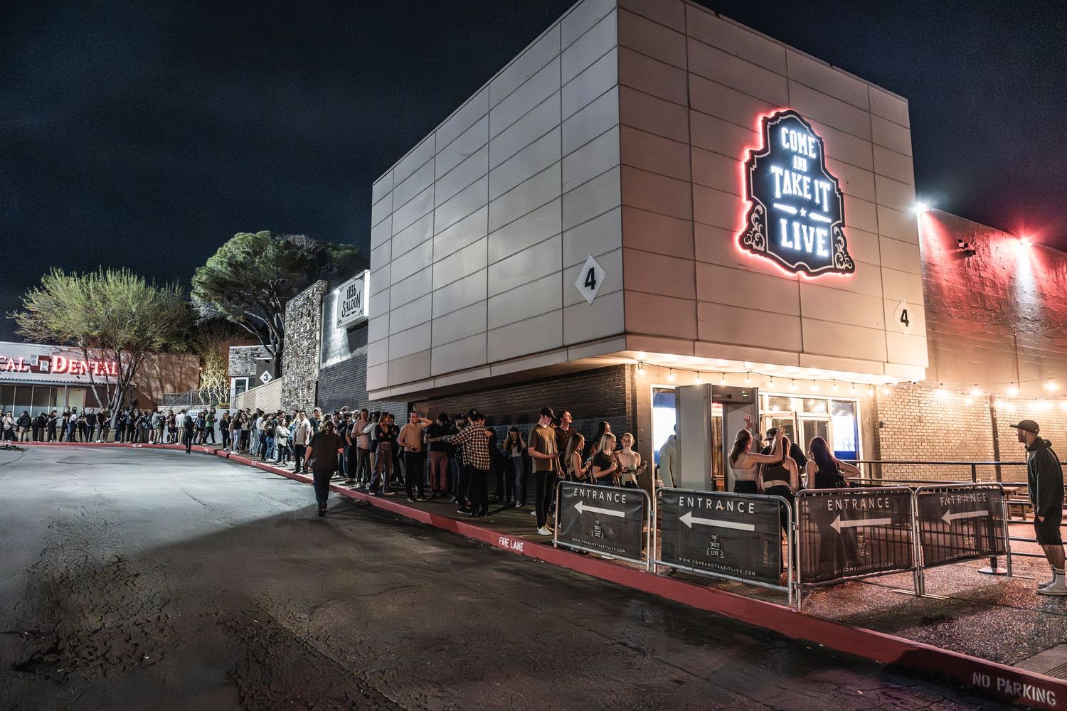 A long line of people waiting outside a music venue at night with illuminated signage.