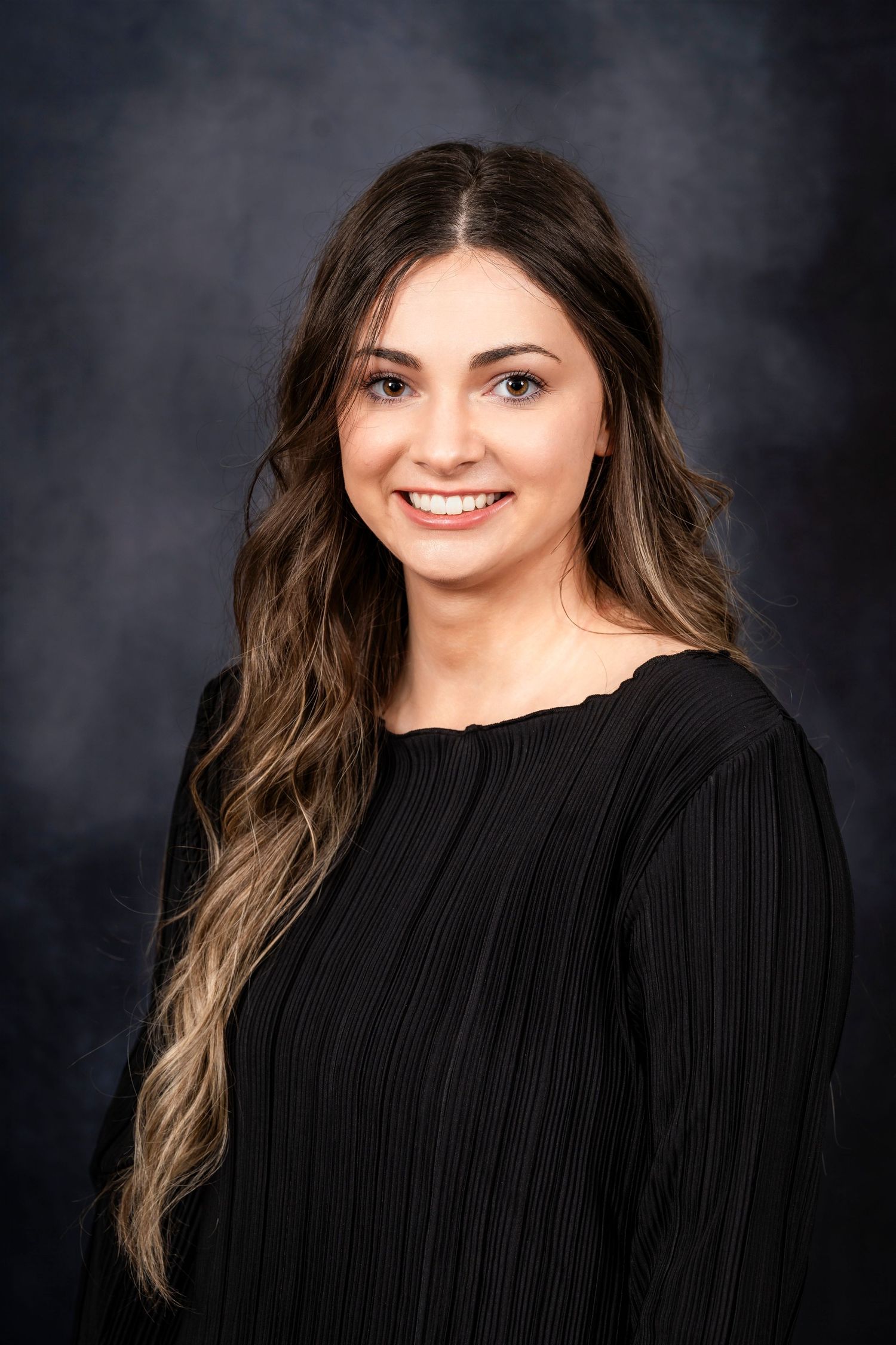 Professional headshot of a woman with long brown ombre hair wearing a black ribbed sweater against a dark background.