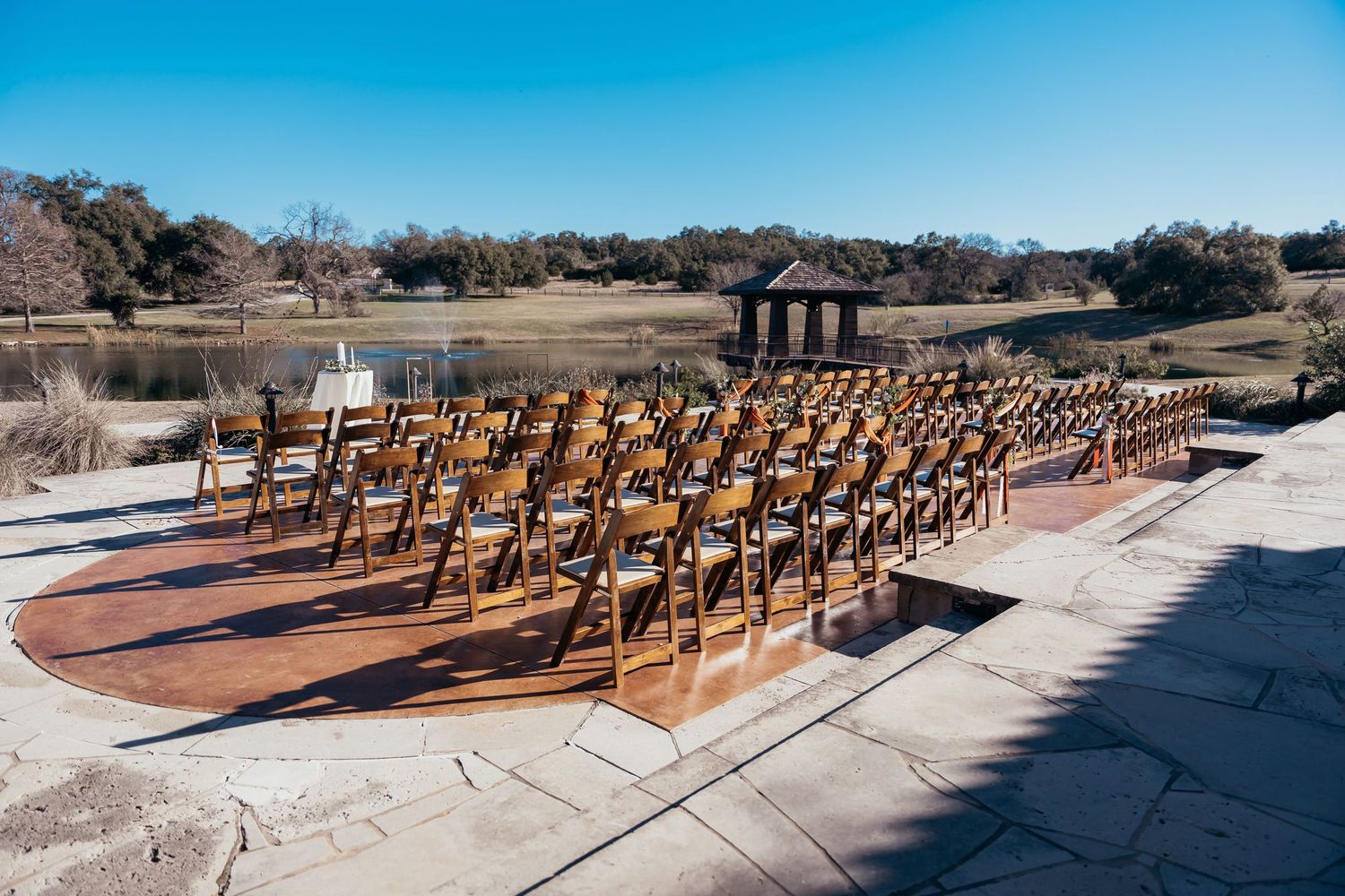 Outdoor wedding ceremony setup with wooden chairs arranged in rows facing a gazebo under bright blue sky.