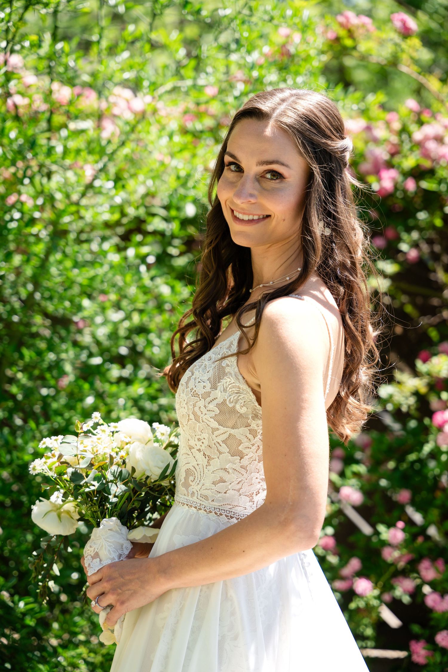 Elegant bridal portrait in a white lace dress against a background of flowering pink bushes.