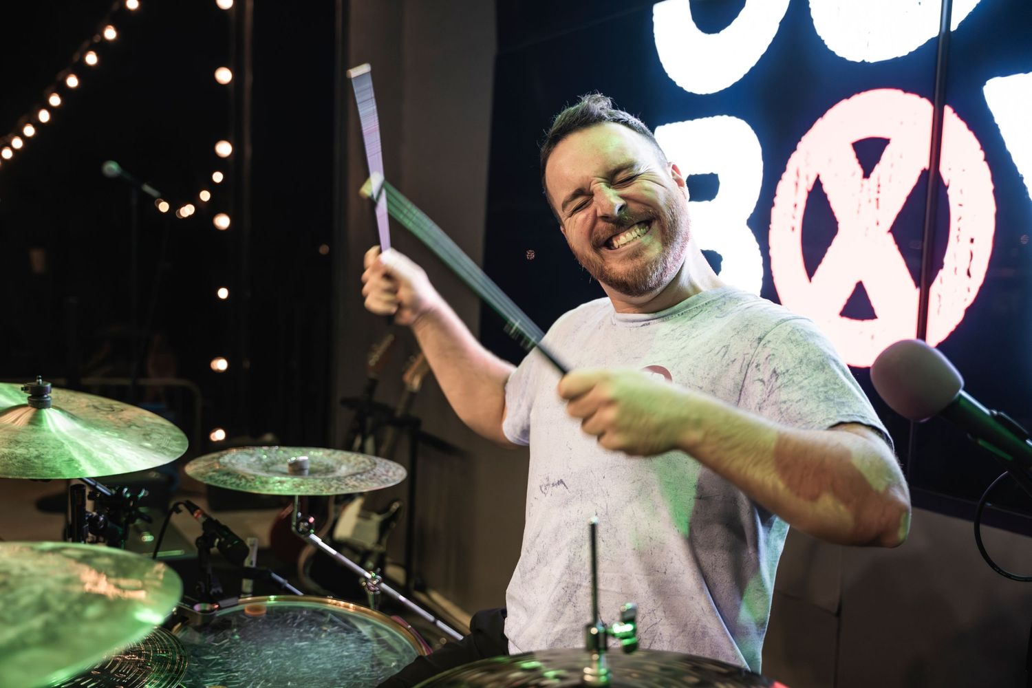 Drummer enthusiastically plays with drumsticks raised during live performance.