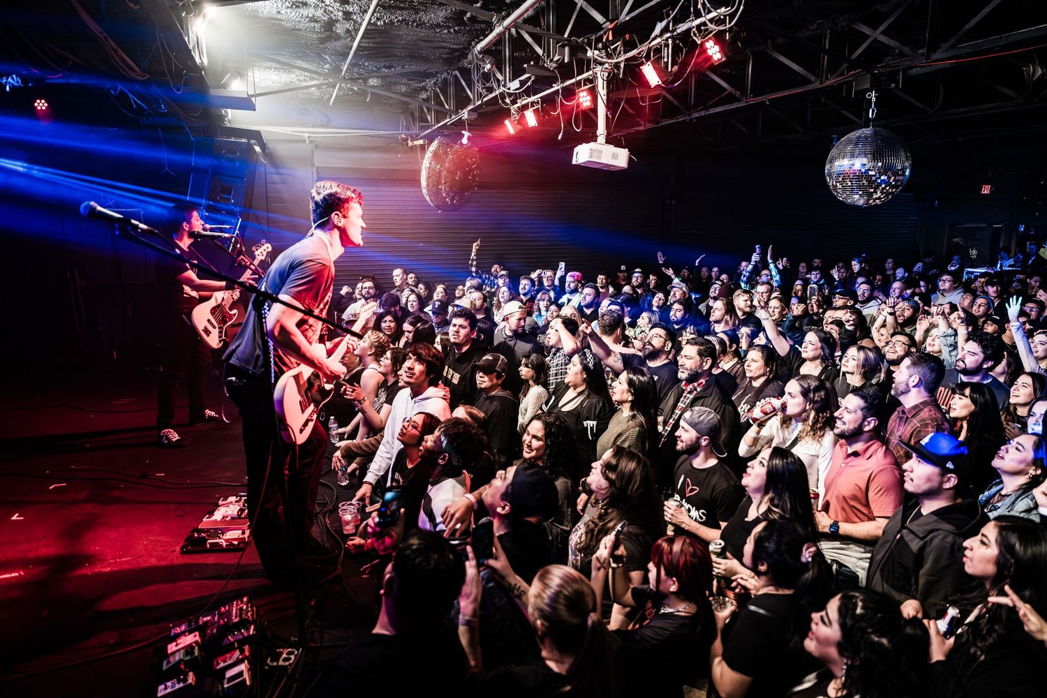 Guitarist plays to enthusiastic crowd under red and blue stage lighting at packed music venue.