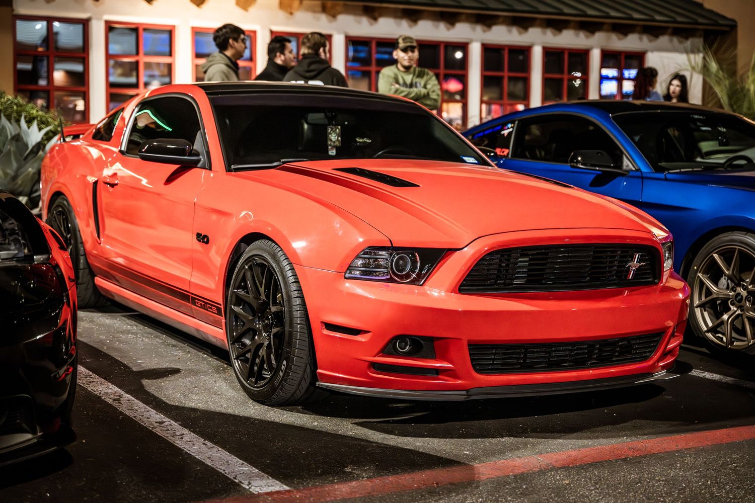 A coral red Ford Mustang GT with black wheels parked at a nighttime car show.