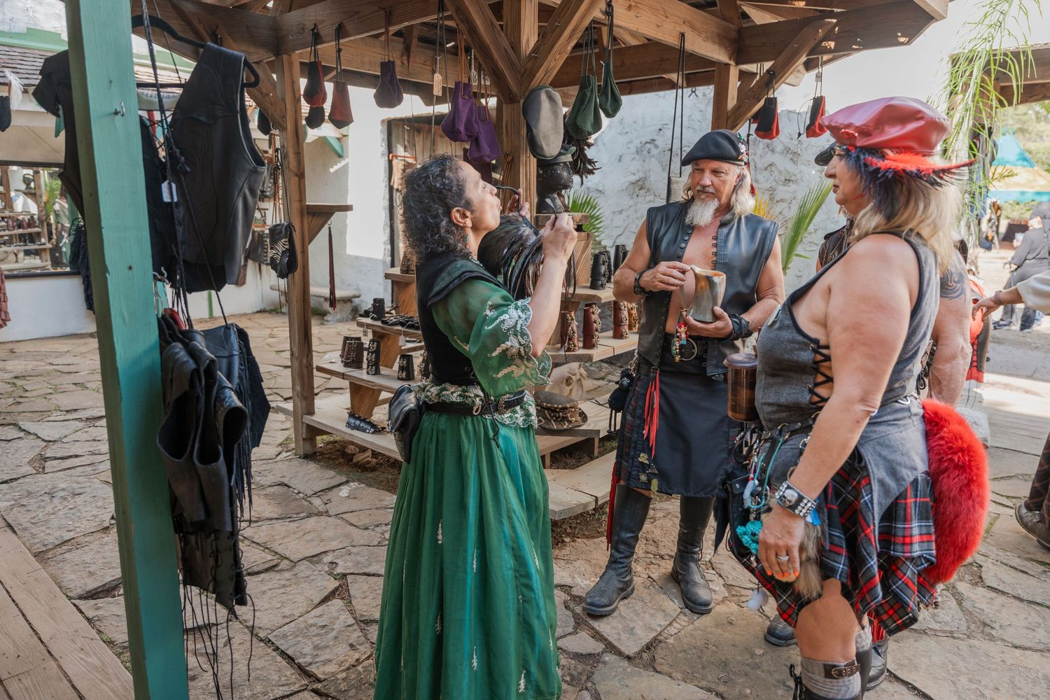 Medieval fair vendors in period costumes interact with customers at an outdoor marketplace.