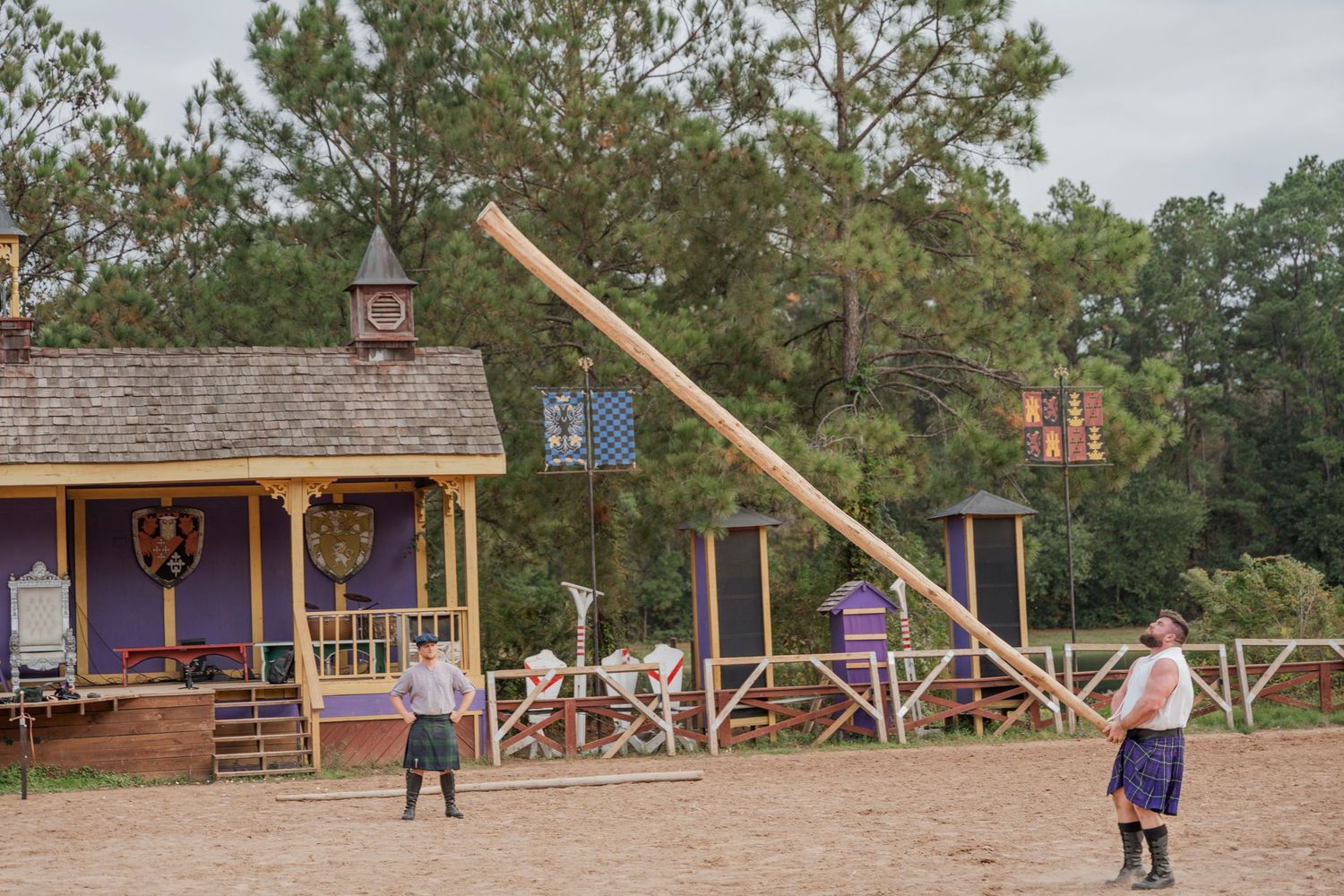 Caber toss demonstration at renaissance faire with traditional wooden buildings visible.