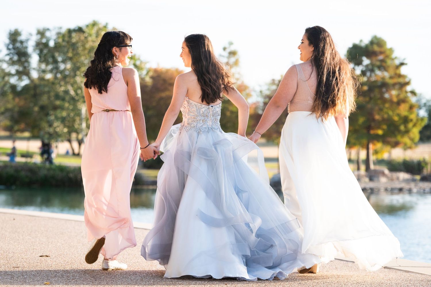 Three friends in formal dresses walk together along waterfront path.
