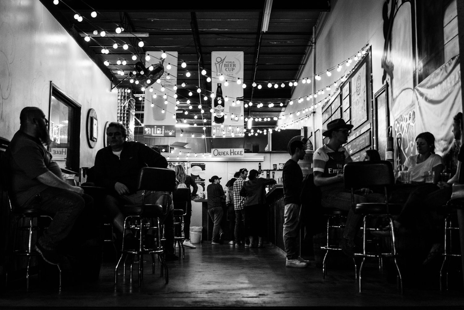 Black and white photo of people sitting at bar counter with hanging string lights creating moody ambiance.