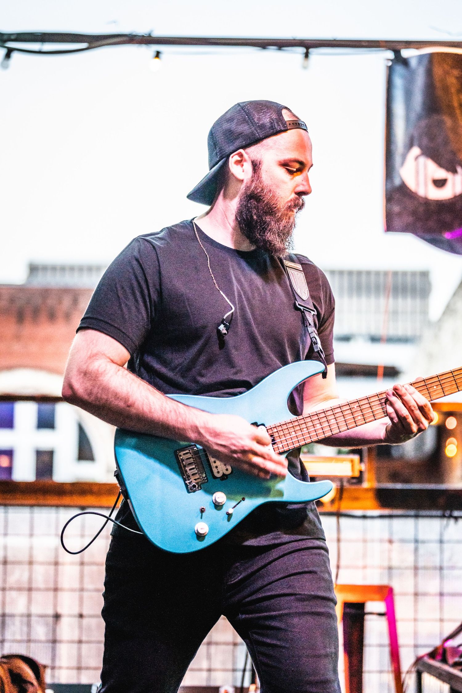 Bearded musician playing blue electric guitar wearing black t-shirt and baseball cap.