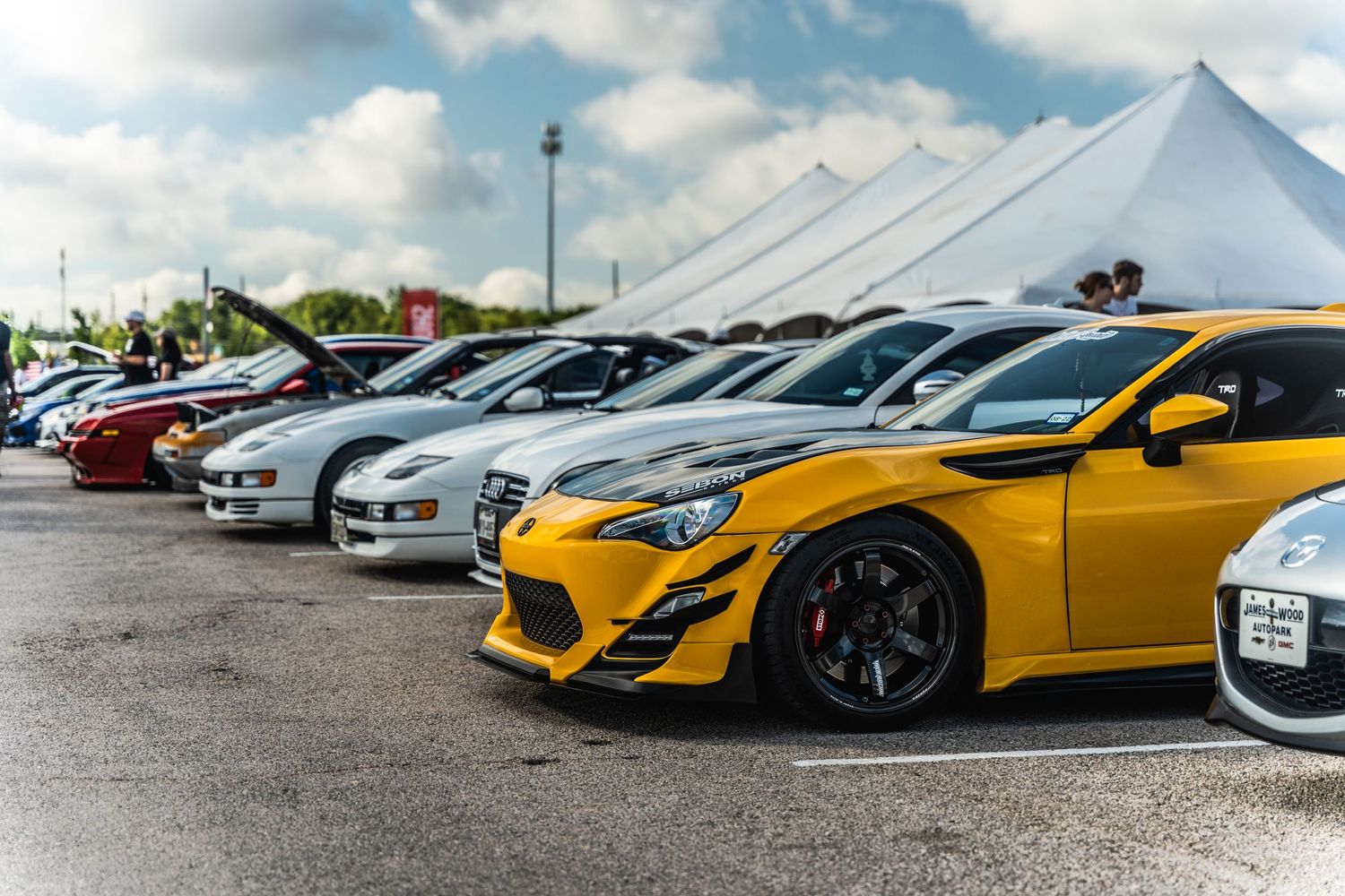 Row of modified cars including a yellow sports car at an outdoor auto show under white tents.