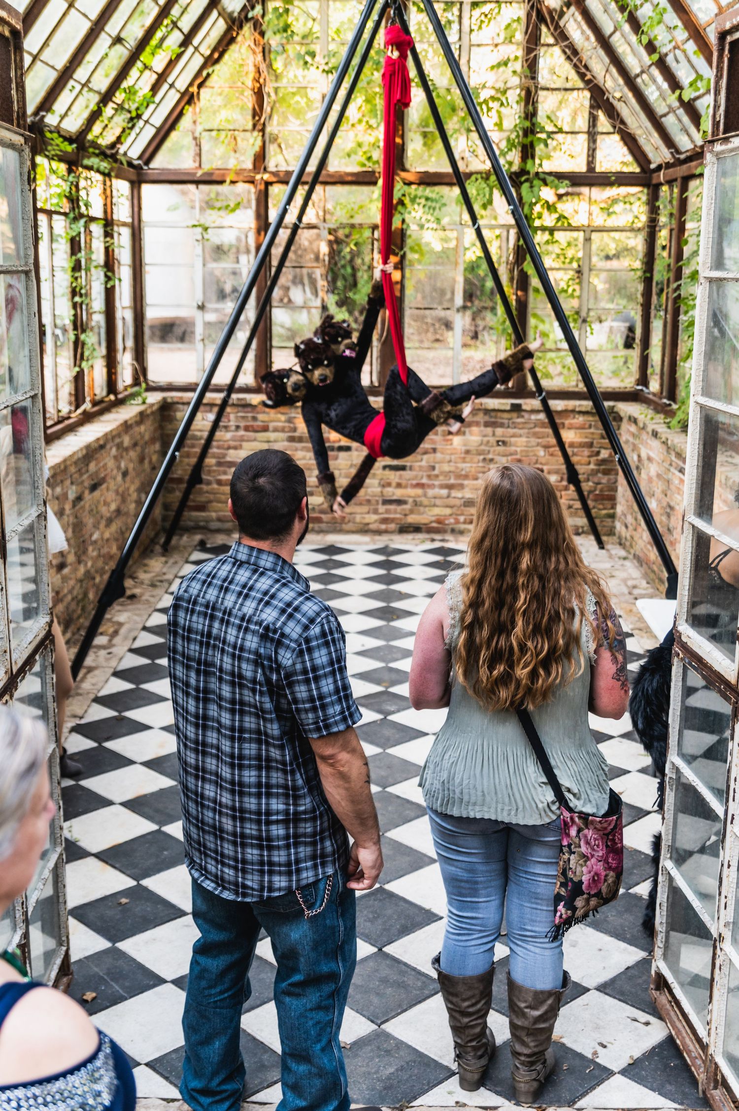 Spectators watch an aerial silk performer in a sunlit conservatory with checkered floor.