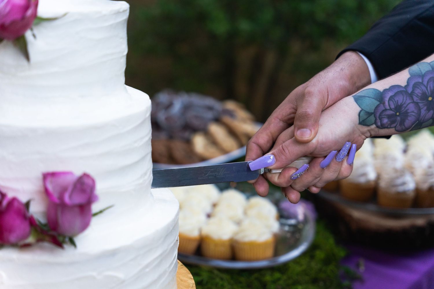 Hands holding a cake knife together to cut into a white wedding cake decorated with purple flowers.