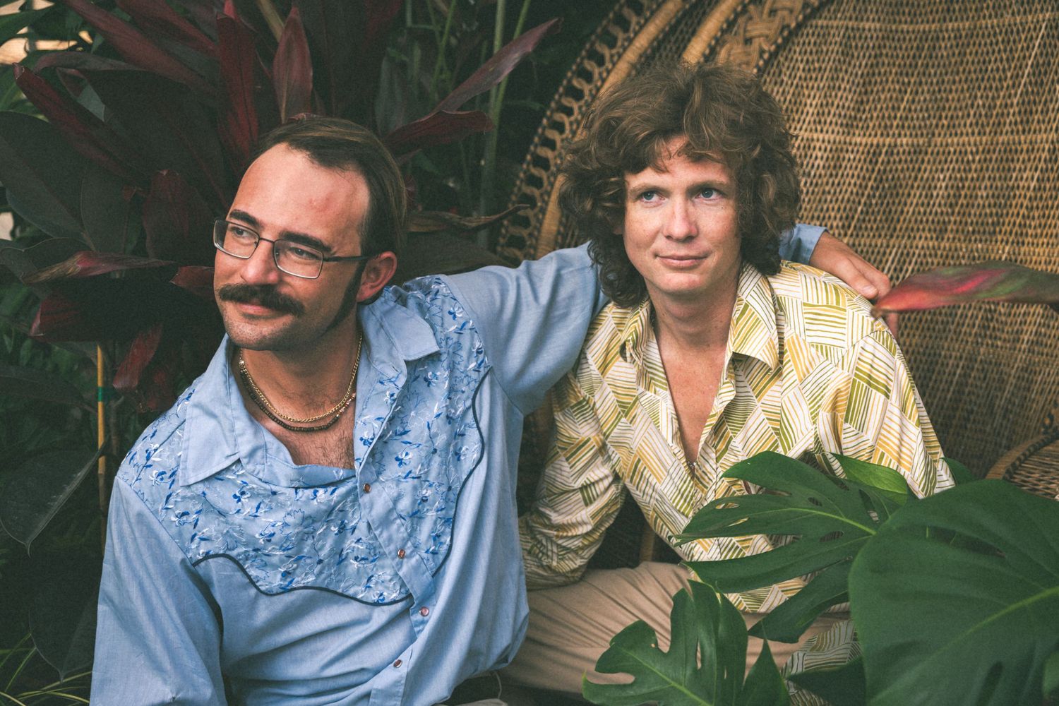 Two people seated on wicker chair surrounded by tropical plants in vintage style photo.