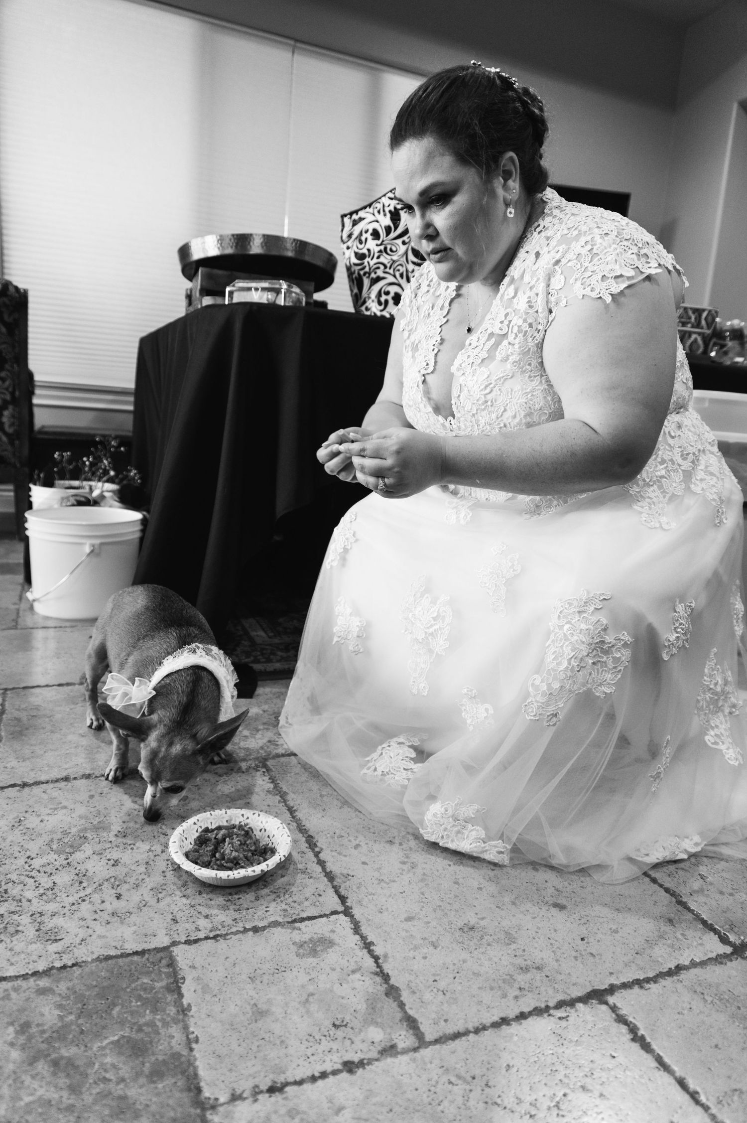 Bride in lace gown sits on floor sharing moment with pet during wedding preparations.