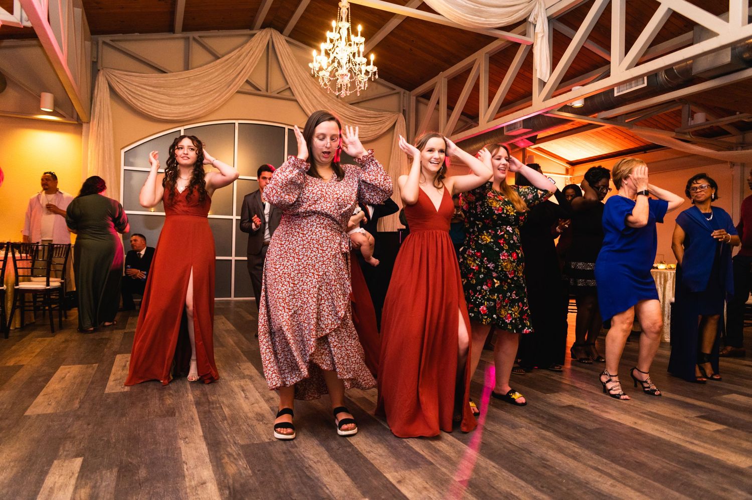 Wedding guests dance together at an indoor reception venue with exposed wooden beams and draped decorations.