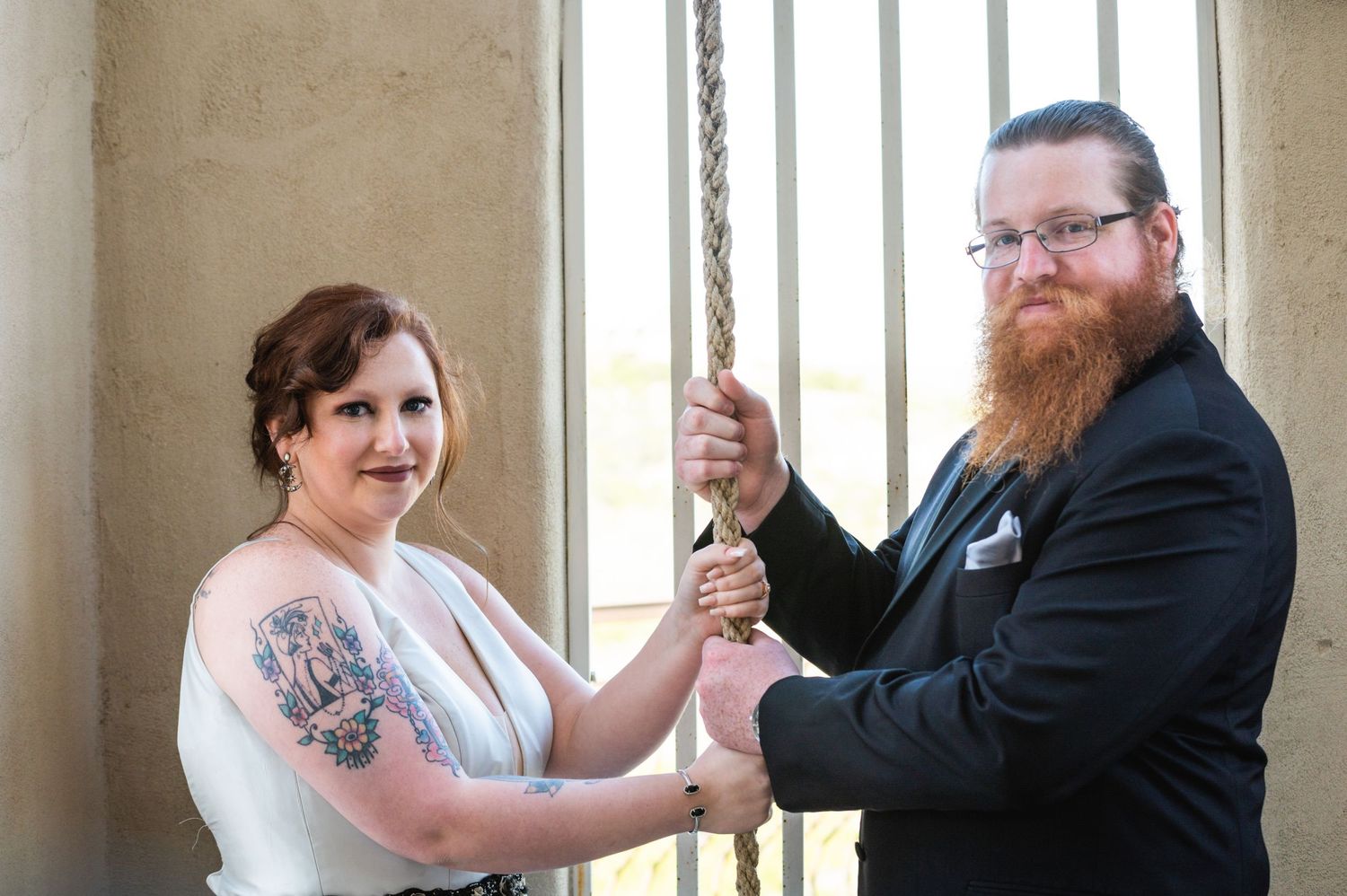 Couple in wedding attire pose while holding a decorative rope together by a window.