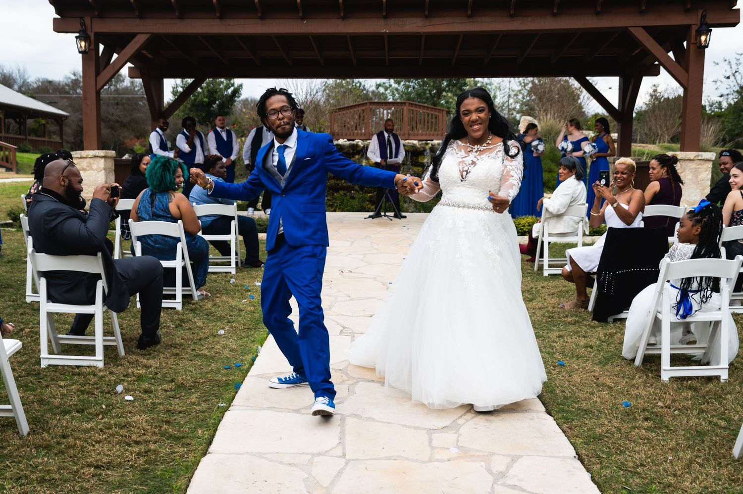 Newlyweds celebrate on stone pathway with wedding guests seated in white chairs.