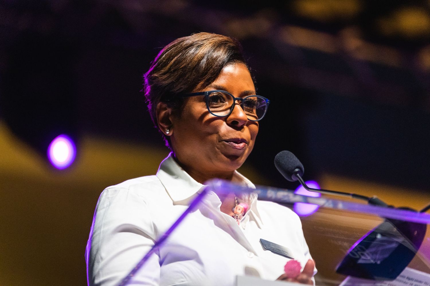 Speaker at podium wearing glasses and white blazer gives presentation with purple stage lighting.