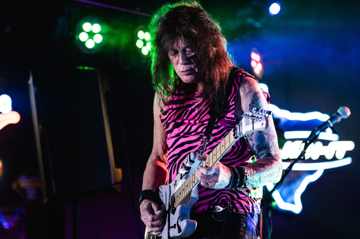 Rock guitarist in pink-lit concert venue plays electric guitar while wearing zebra print.