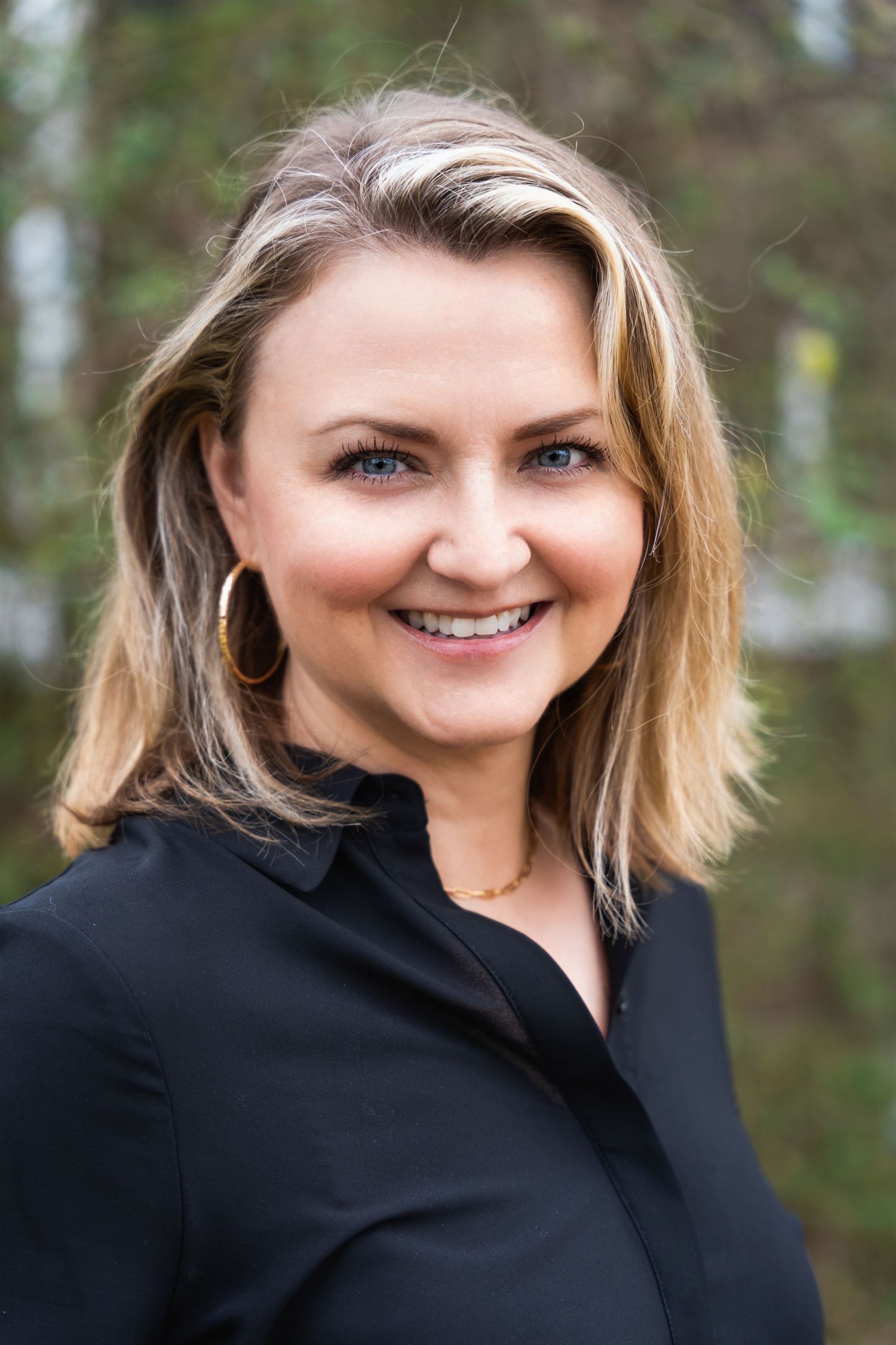 Business portrait of person in black top with shoulder-length blonde hair against outdoor setting.