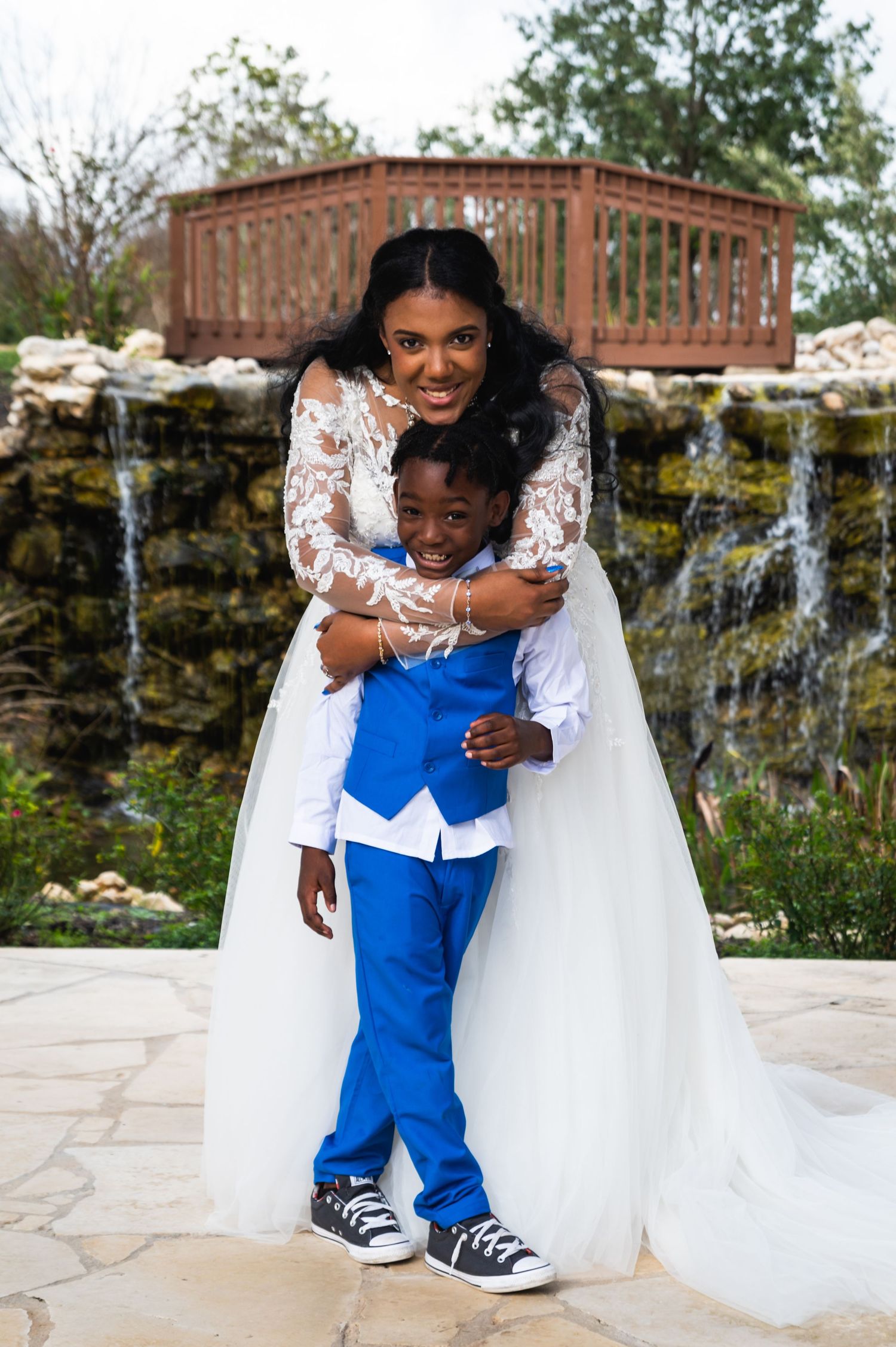 A tender moment captured between a bride in white lace and small attendant in bright blue at an outdoor venue.