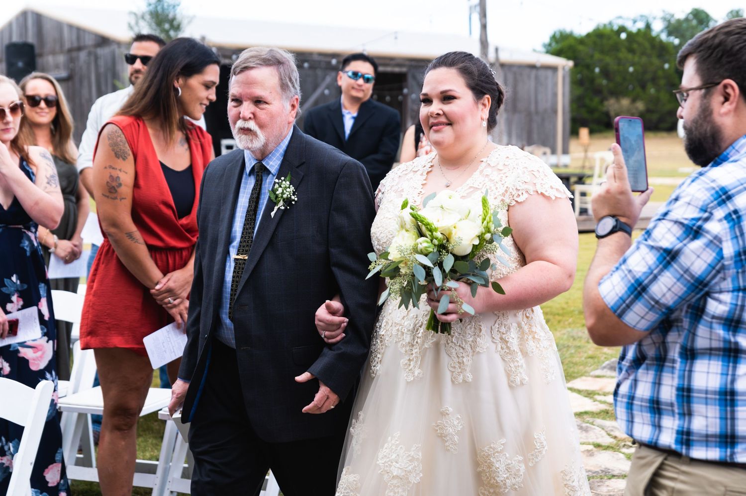 A joyful wedding procession walks along an outdoor path with guests looking on.