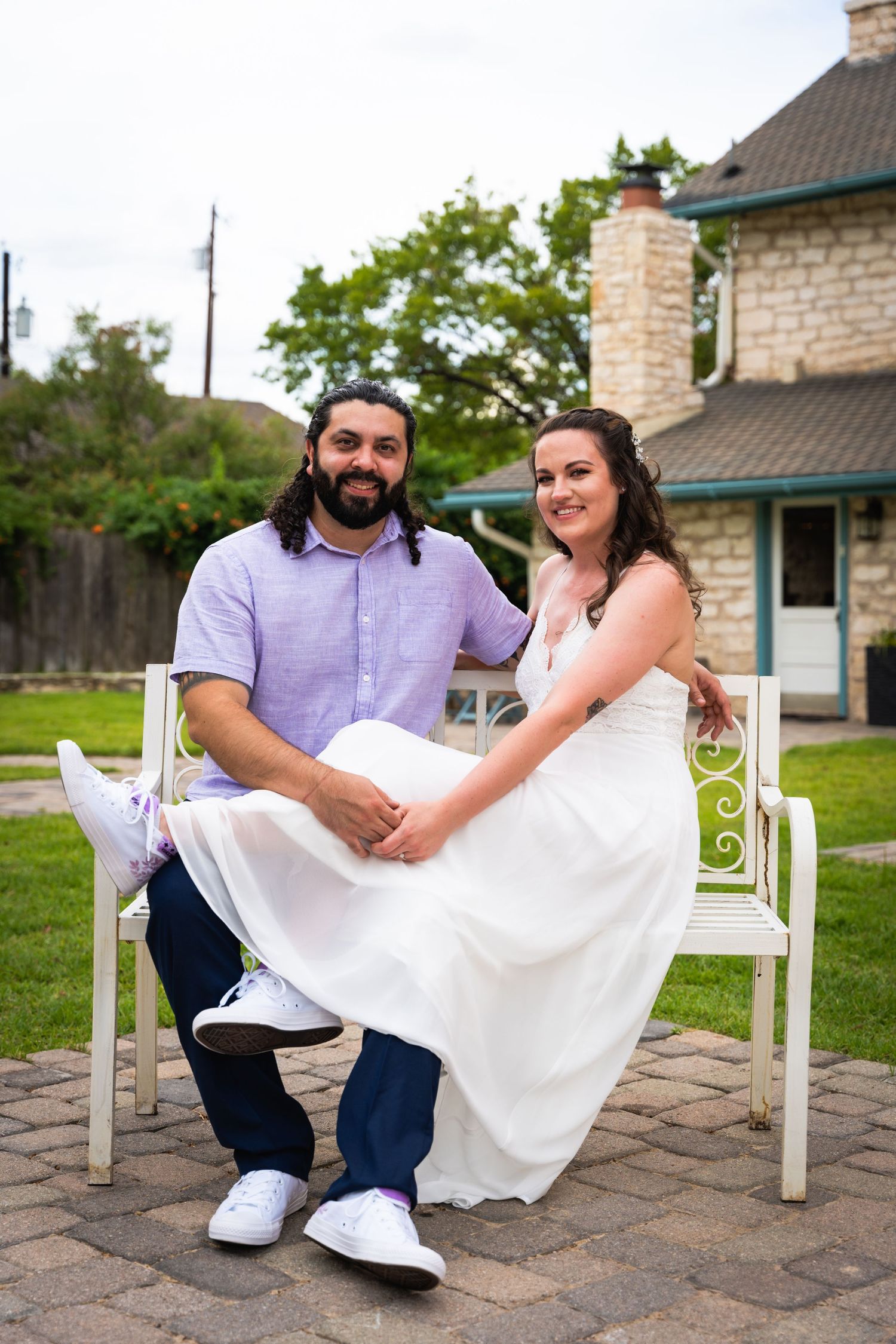 Couple sitting together on white bench in garden setting wearing wedding attire.