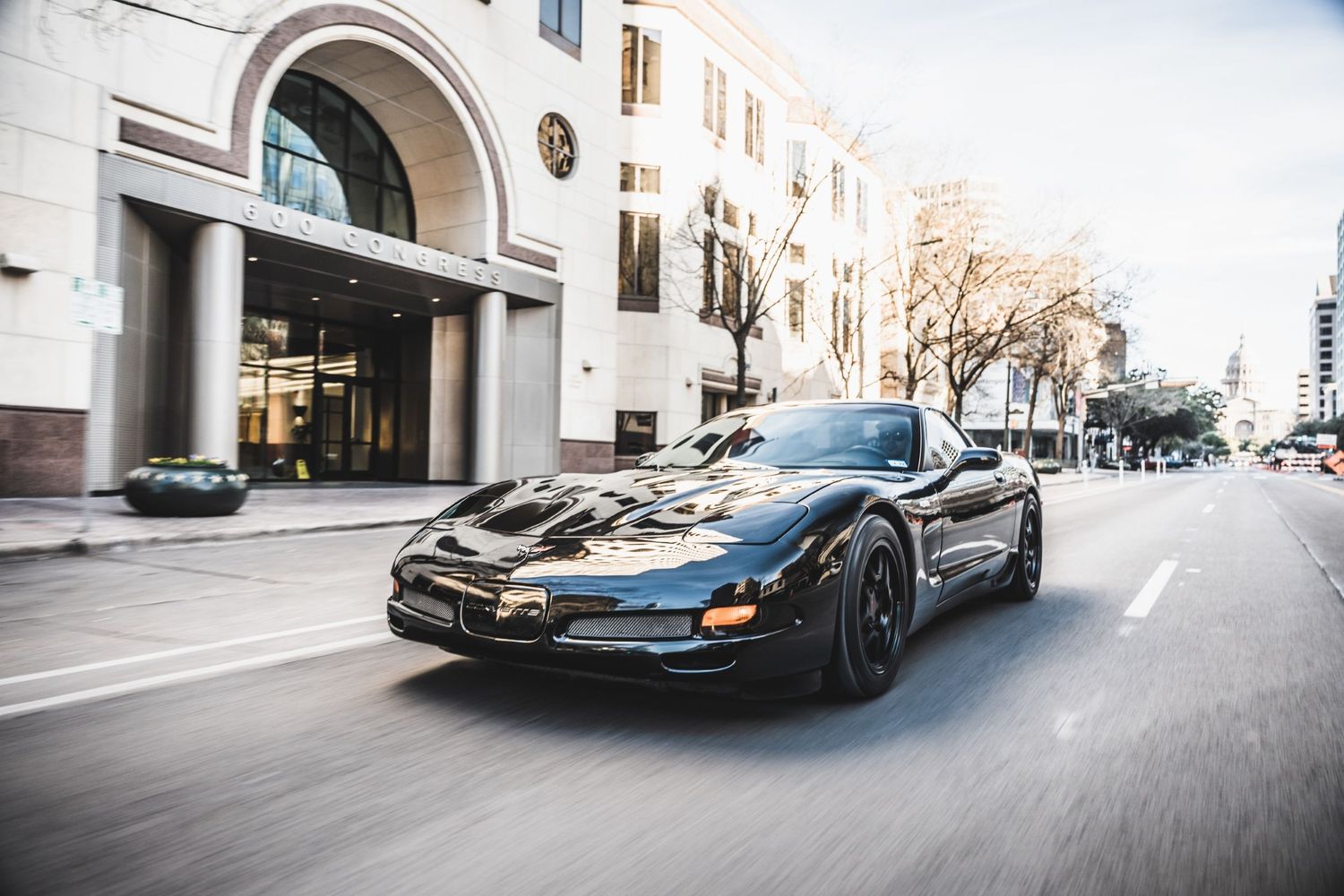 Black sports car with sleek aerodynamic modifications cruising on an urban street with classical architecture.