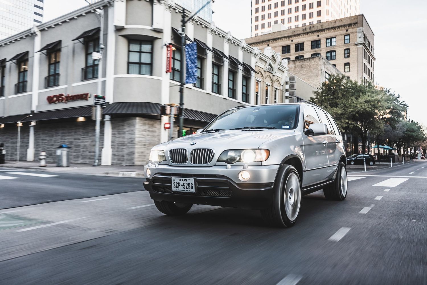 Silver BMW X5 SUV driving on city street with urban architecture in background.